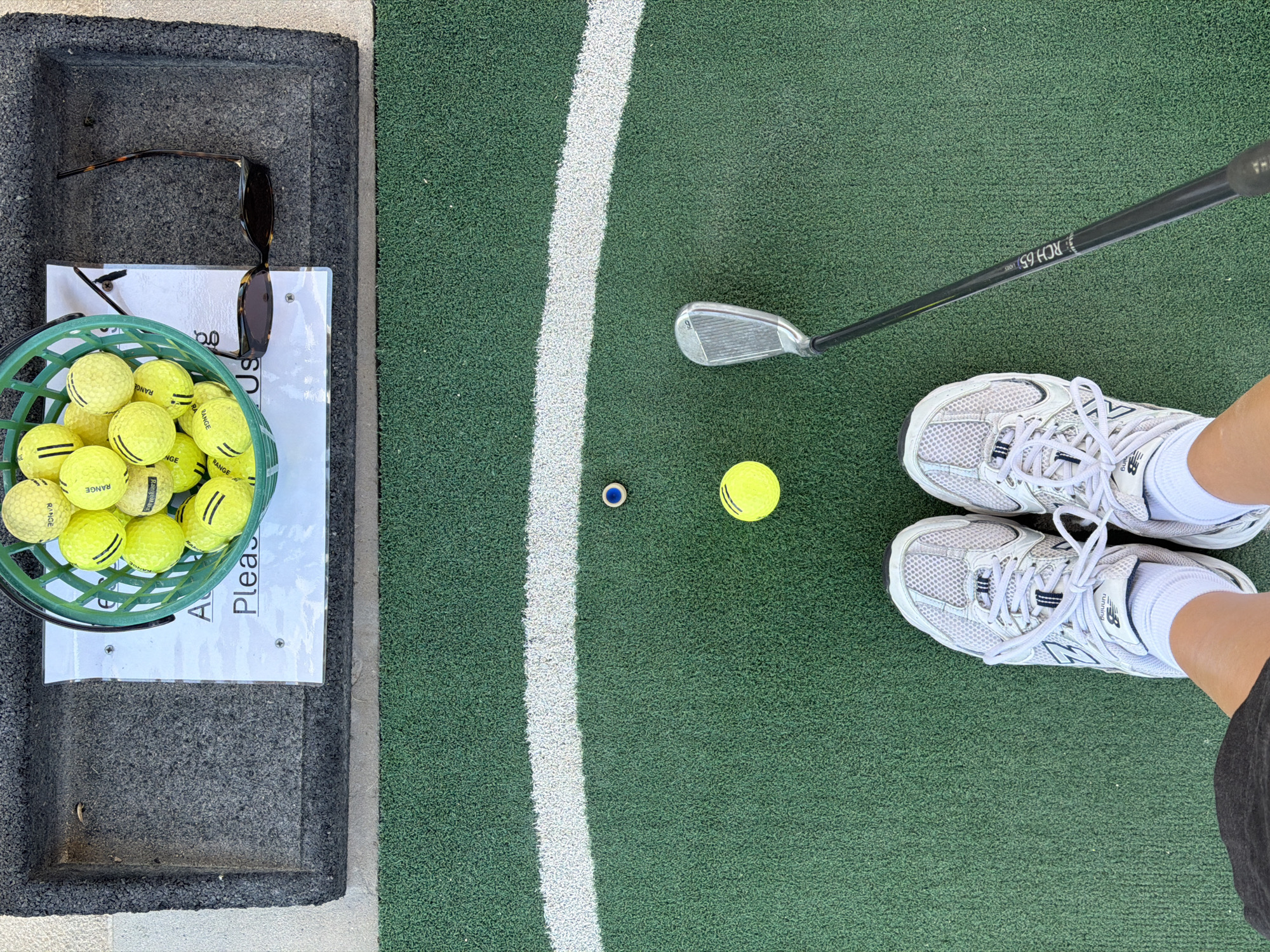An overhead shot shows a person's feet wearing white New Balance running shoes on a green artificial turf golf practice area. A golf club, identified by the text "RCH 65", is positioned to the left of the frame, with its head near the turf. Two yellow golf balls are visible on the turf, one closer to the club and the other further away. A white line curves across the turf in the mid-ground. In the foreground, a dark mat contains a pair of sunglasses and a green plastic basket filled with yellow golf balls. A white sign with text is also present on the mat. The location is a golf driving range.