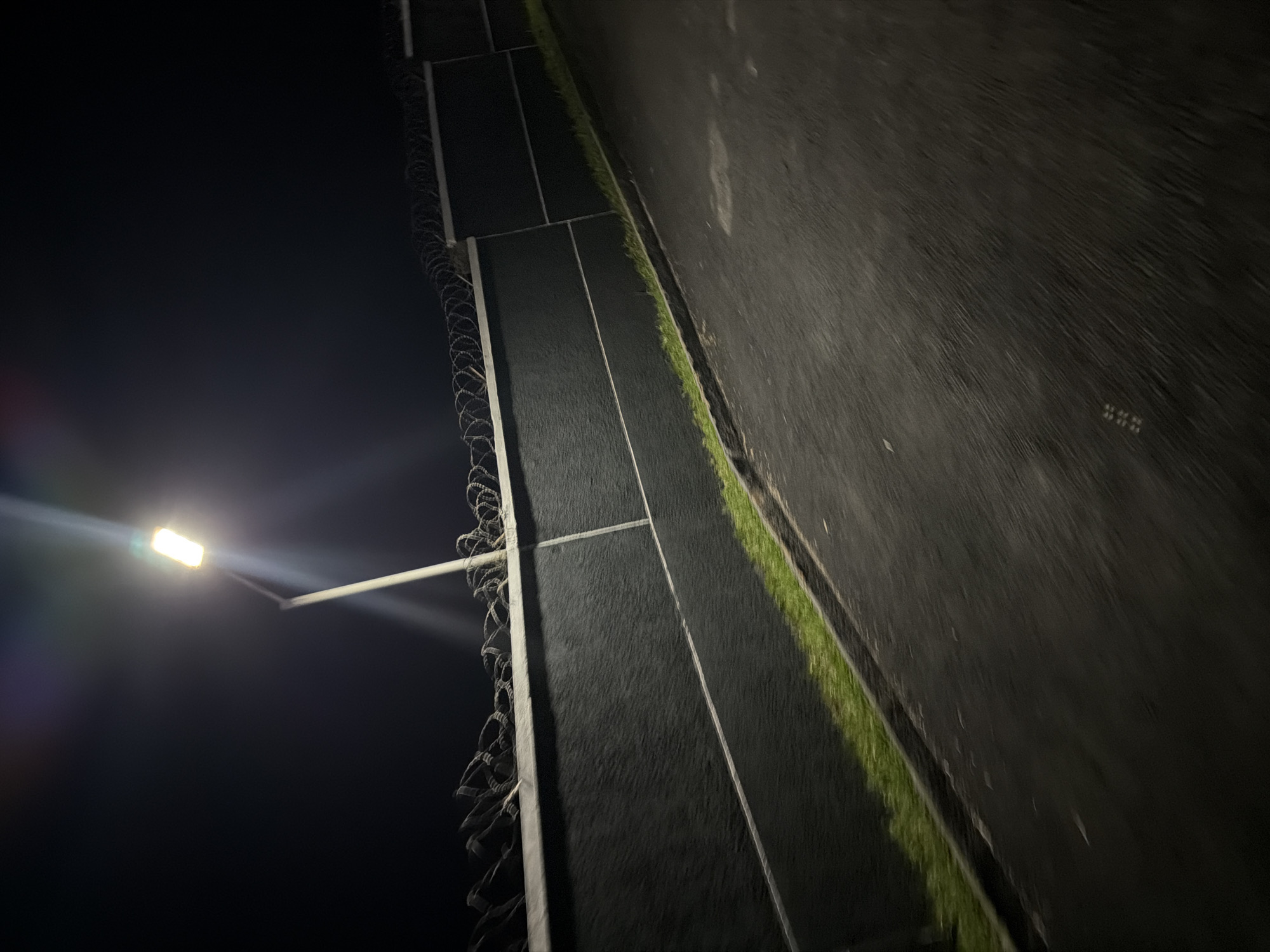 A security light illuminates a dark night scene in Bukuru, Nigeria, casting a bright glow on a tall, dark wall topped with barbed wire. The wall, which appears to be constructed of concrete or a similar material, is segmented by vertical lines. Adjacent to the wall is a strip of green grass, and beyond that, a paved road is visible. The image is slightly blurred, suggesting it may have been taken from a moving vehicle. The overall impression is one of a fortified or secured area, with the harsh lighting emphasizing its perimeter and the absence of any visible people or activity.