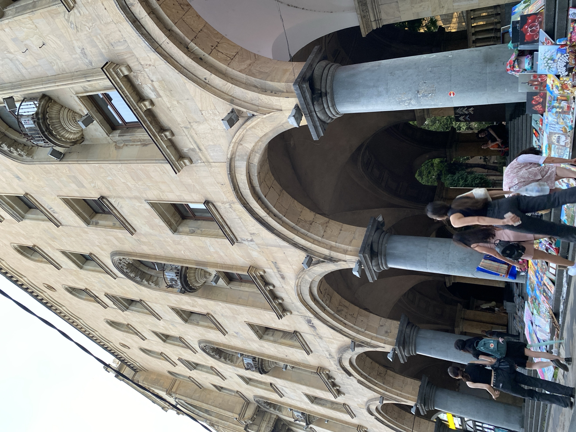 The image depicts a street scene in Tbilisi, Georgia, focusing on a grand, multi-story building with a beige stone facade, characterized by arched windows, balconies, and colonnades. The perspective is from below, looking upwards at the architecture, which exhibits classical and possibly Art Nouveau influences. A prominent feature is the series of large, rounded arches forming a covered walkway at the ground level. Several tall, grey cylindrical columns support the arches.
In the foreground, several people are visible. Two women in dark clothing are walking away from the camera, towards the left. One has a backpack. A third woman in a pink dress, also with a backpack, is walking in the same direction. A fourth person, wearing a patterned dress, is bending over to examine art displayed on the ground. The art appears to be paintings, some with still-life subjects like fruit and bottles, laid out on the steps and pavement in front of the building. Further back, under the arches, a couple is seated on steps, with greenery visible behind them.
The environment suggests an outdoor marketplace or a pedestrian area in a historical part of the city. The lighting indicates it is daytime, possibly late afternoon, given the soft light and shadows. The overall impression is one of a vibrant urban setting with a blend of historical architecture and contemporary street life. The building's design, with its detailed ornamentation and stone construction, points to a significant historical structure, possibly a government building or a grand residential complex.