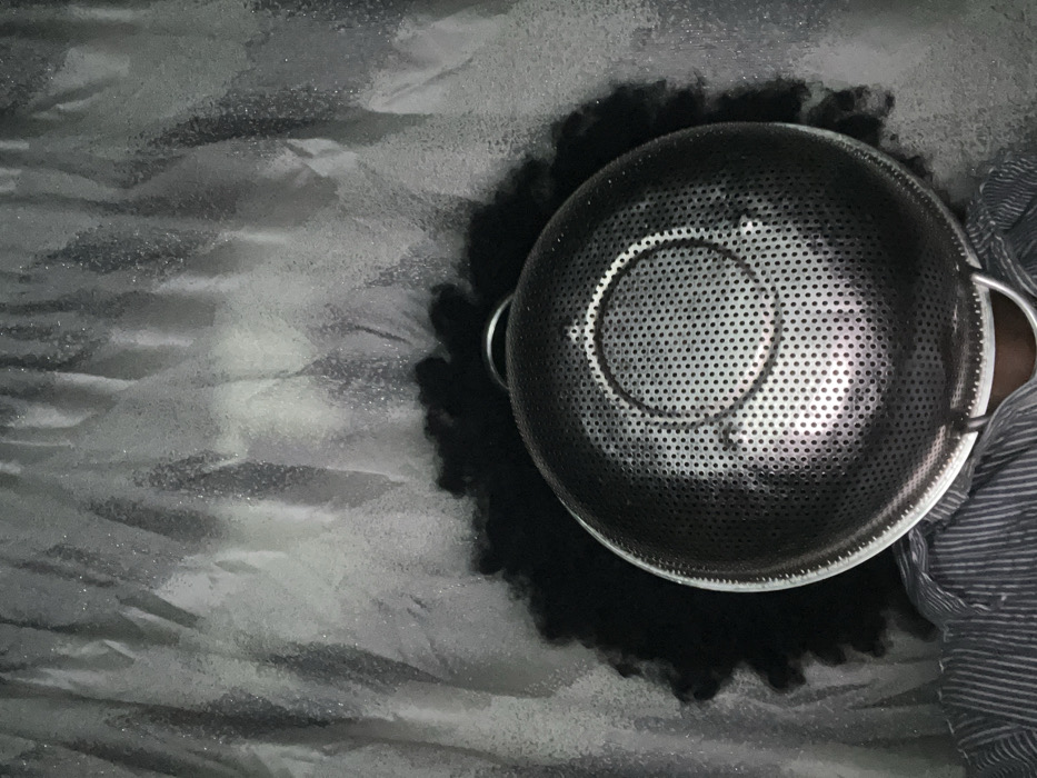The image depicts an individual in Iranla, Nigeria, whose face is completely obscured by a large, circular, perforated metallic colander. The colander, appearing to be stainless steel, features numerous small, uniformly distributed holes and a concentric ring pattern on its interior base. Two metallic wire handles are visible on opposite sides of the colander's rim, with one partially visible at the bottom. Dark, curly, voluminous hair is visible around the upper and side edges of the colander. The individual is wearing a light-colored shirt with horizontal stripes, visible on the lower left. The background consists of a textured, light grey material, likely fabric or a curtain, displaying vertical folds and a subtle reflective sheen.