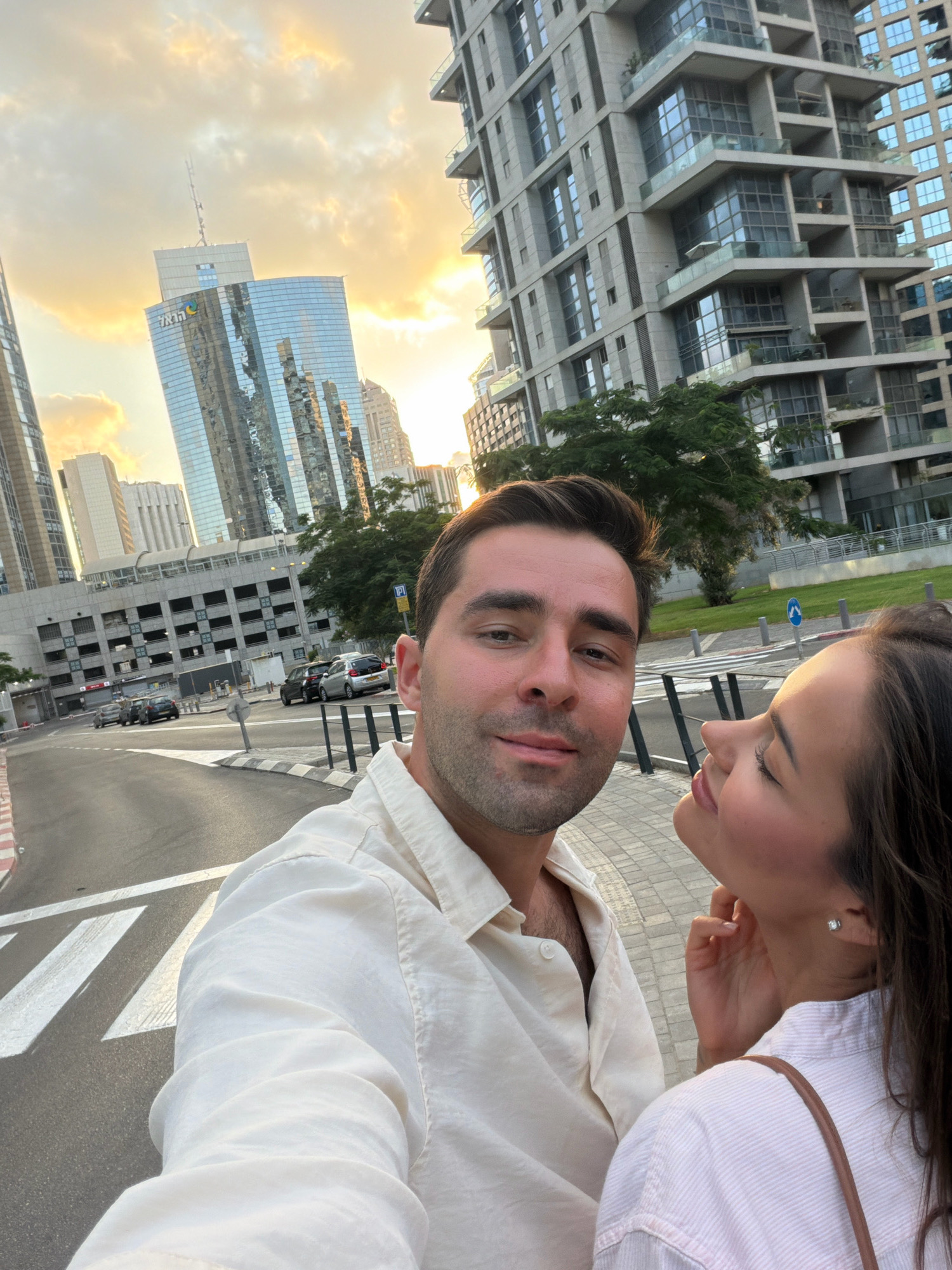 A selfie taken by a man and woman in Ramat Gan, Israel. The man is in the foreground, wearing a white button-down shirt. The woman is to his right, her head tilted up, with her hand touching her neck. Behind them, modern skyscrapers rise against a cloudy sky with golden sunlight. A street with crosswalks and parked cars is visible in the mid-ground, with a multi-story parking garage and trees bordering it. The overall scene is an urban environment during what appears to be sunset or sunrise.