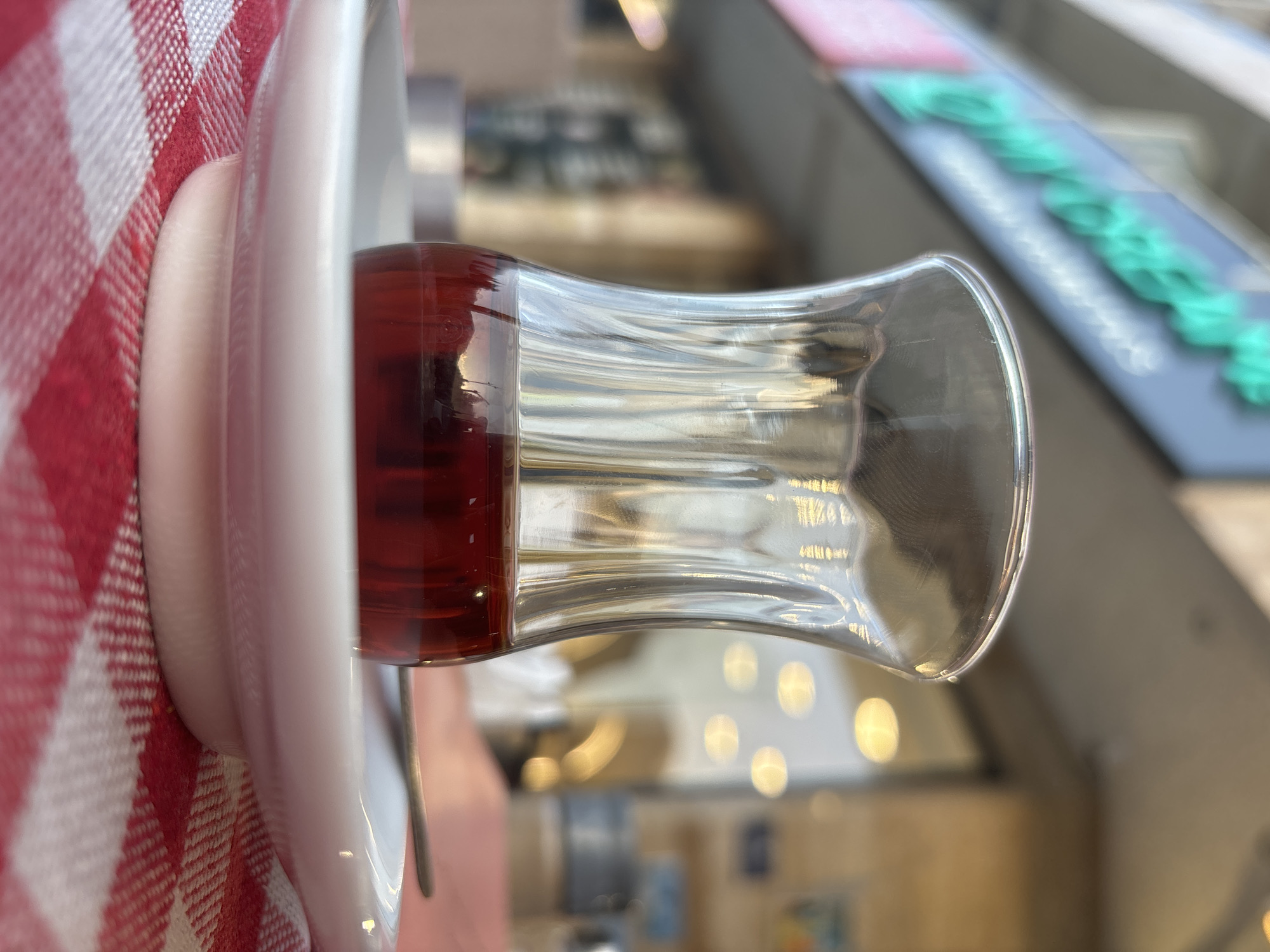 A close-up shot focuses on a clear glass filled with dark red liquid, likely tea, resting on a white saucer. The saucer is positioned on a red and white checkered tablecloth. In the background, a blurred urban scene is visible, featuring a building with illuminated signage in bright green. The focus is sharp on the glass and saucer, with the background softly out of focus, creating a shallow depth of field. The overall lighting suggests an outdoor setting, possibly a cafe or restaurant in Budva, Montenegro, during daylight. There are no people or discernible actions visible in the image.