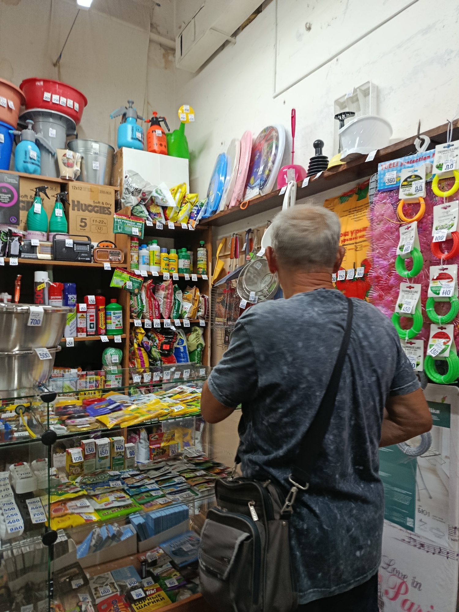 A man with gray hair is standing in a cluttered store, facing away from the camera. He is wearing a dark gray t-shirt and has a dark shoulder bag. The store appears to be a small, independent shop, possibly selling household goods or hardware. Shelves are packed with various items, including cleaning supplies, gardening tools, and packaged goods. Some products are displayed in plastic bins on glass shelves.
Visible text includes brand names like "HUGGIES" and product descriptions in Russian, such as "ДИХЛОФОС" (Dichlorvos, an insecticide), "КОМПОСТ" (Compost), and "БОРНЫЕ ШАРИКИ" (Boron balls, likely referring to rodent poison). Prices are also visible on many items, with numbers like "150", "450", and "700". The overall impression is of a well-stocked, somewhat disorganized store. The setting appears to be indoors, and there are no clear indicators of the time of day or weather. The scene suggests a casual shopping experience.