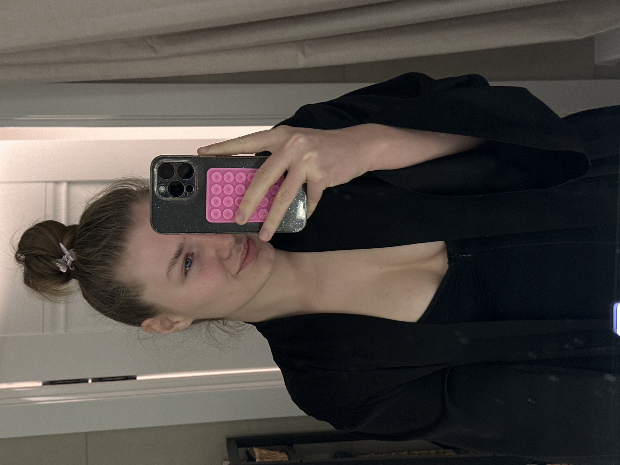 A person with light skin and hair pulled into a bun is taking a selfie in a bathroom. The person is wearing a black robe or jacket over a black top. The phone has a glittery case with a pink pop socket. The background shows a bathroom with a white door and beige walls. There is a curtain or blind partially visible above the door frame. The photo shows the reflection in a mirror with a small amount of the surroundings visible. The location is most likely indoors in a private residence. There is no information in the image to suggest the city of Kazan, Russia.