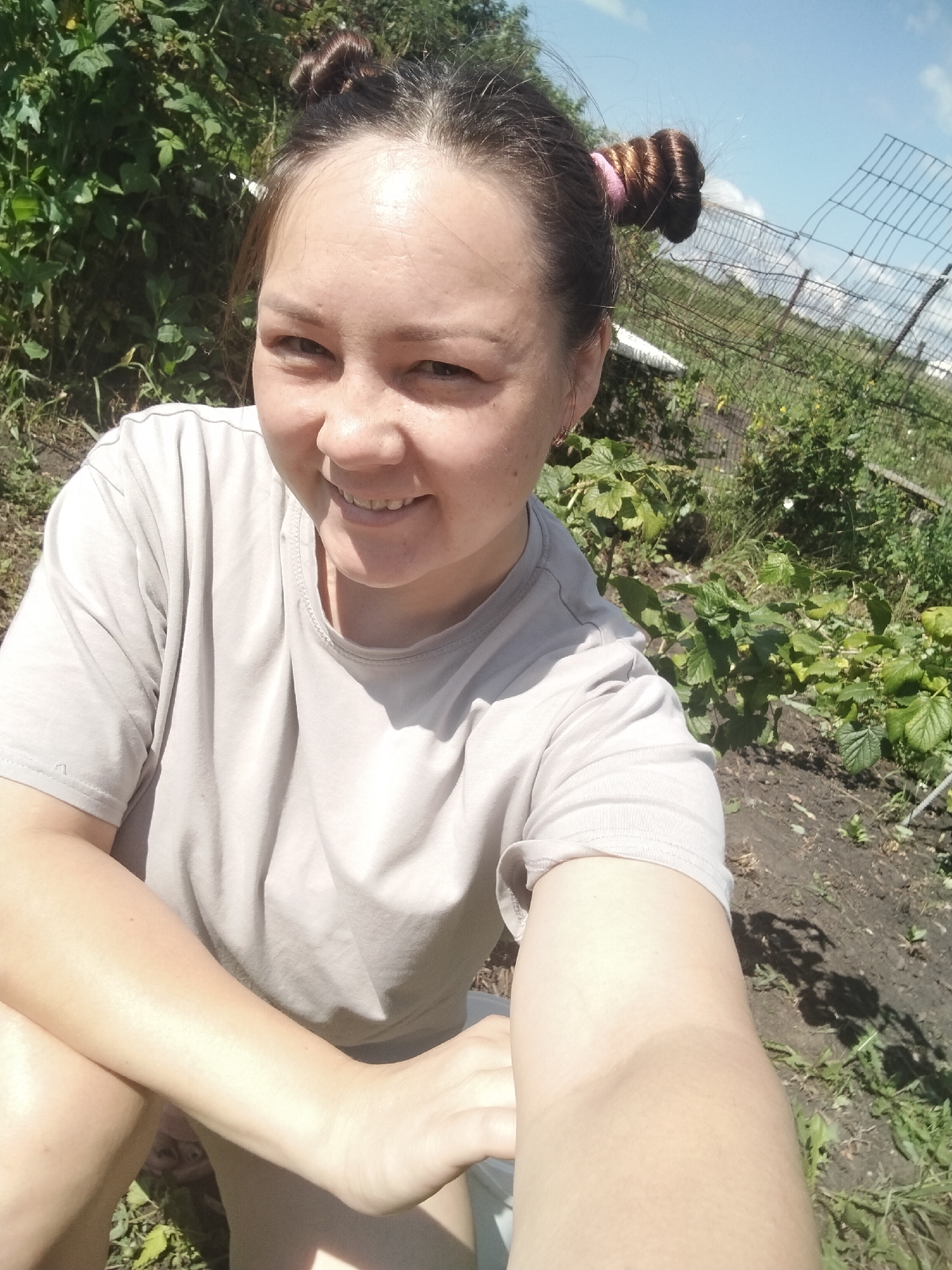 In the city of Vazherki, Russia, a young woman with dark hair styled in two buns is taking a selfie outdoors. She is wearing a light gray t-shirt and is seated or squatting in a garden with green foliage and exposed soil. A wire fence is visible in the background against a bright blue sky. The woman is smiling and looking directly at the camera.