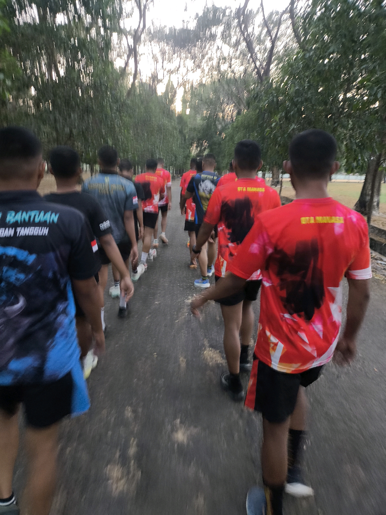 The image depicts a group of individuals, likely males, from behind, walking or jogging on an asphalt path lined with trees. They are wearing athletic attire, mostly t-shirts and shorts, in various colors including red, blue, and black. Some t-shirts have visible text and graphics. The environment appears to be an outdoor park or recreational area with a treeline on either side of the path. The lighting suggests it might be early morning or late afternoon, with sunlight filtering through the trees. The overall impression is of a group engaged in physical activity in a natural setting within a city.