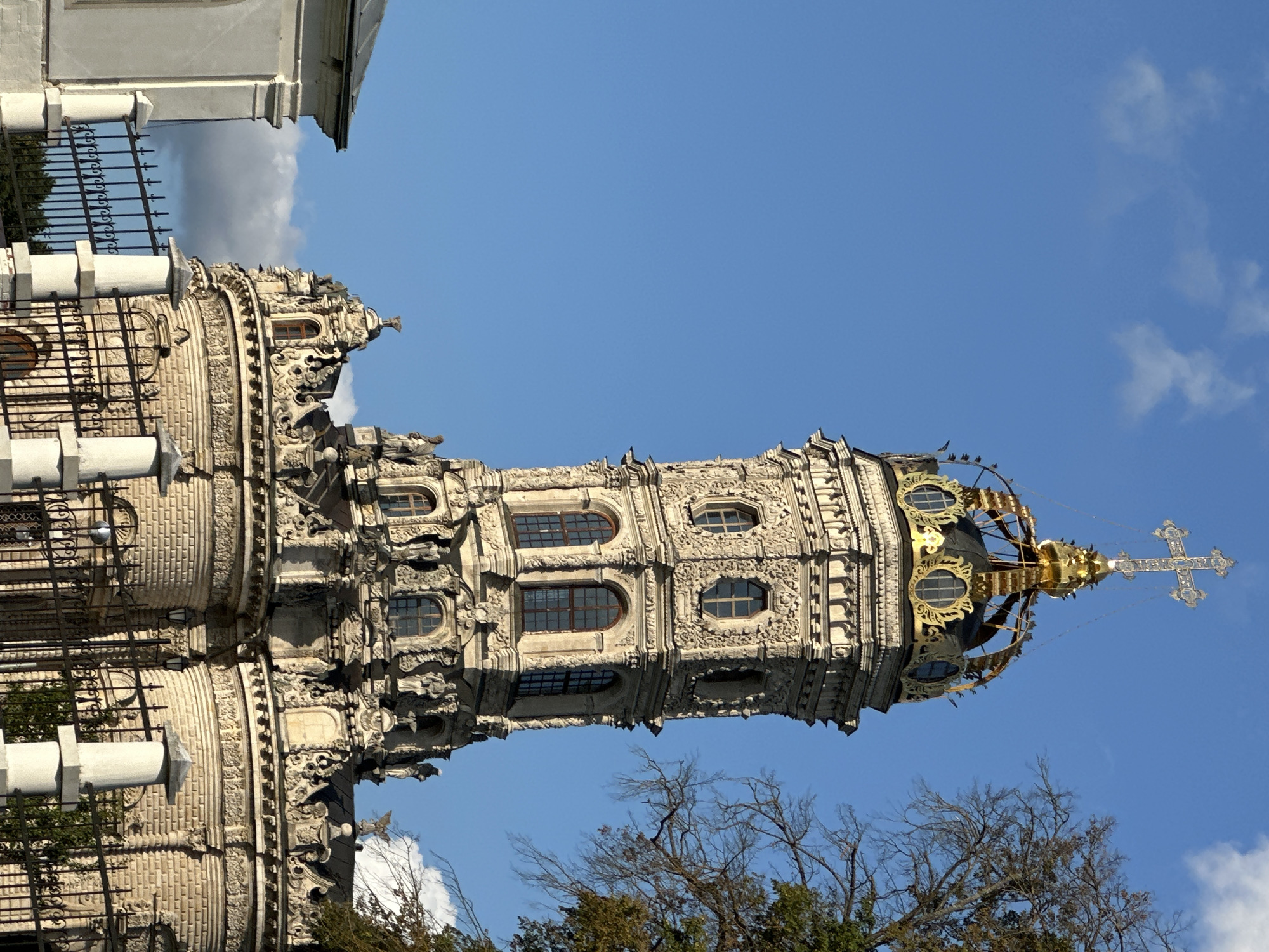 The image shows the Church of the Sign of the Most Holy Mother of God in Dubrovitsy, Russia, under a clear blue sky with a few scattered clouds. The ornate Baroque-style church, made of white stone, is characterized by its elaborate decorations and a distinctive golden crown-shaped dome topped with a cross. The tower is adorned with numerous sculptures and intricate carvings. In the foreground, a black wrought-iron fence with white stone pillars separates the viewer from the church. To the left, a portion of another white building with a slanted roof is visible. A tree with bare branches and some autumn foliage is partially visible on the right side of the frame, adding to the sense of the season. The lighting suggests it is a sunny day, casting distinct shadows on the architectural elements.