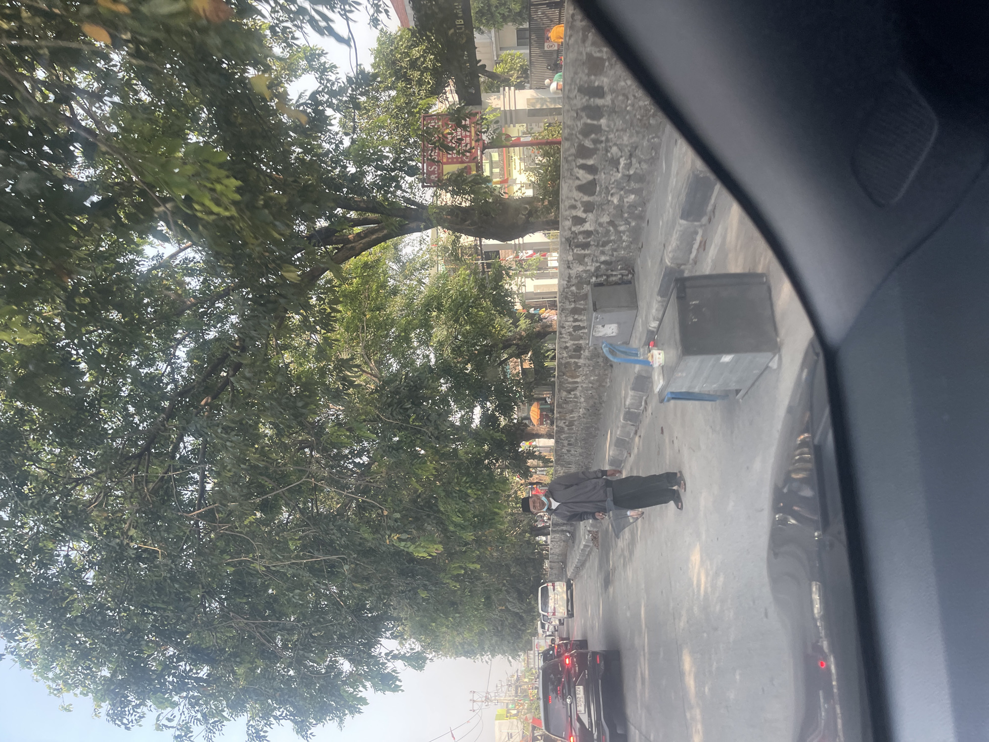 An outdoor daytime scene on a street in Bekasi, Indonesia shows a man standing by the roadside. The man is wearing a grey jacket, dark pants, a light-colored face mask, and a black cap. He is holding a black bag with a plastic bag inside, possibly containing fish. He is standing on the asphalt road next to a concrete curb and wall. Behind him, there is a line of traffic with several cars. Large trees with green foliage line the street, casting shadows. A building with a sign that reads "R. S. S..." is visible in the background, along with other structures and some parked vehicles. The foreground shows the interior of a vehicle, suggesting the photo was taken from a car.