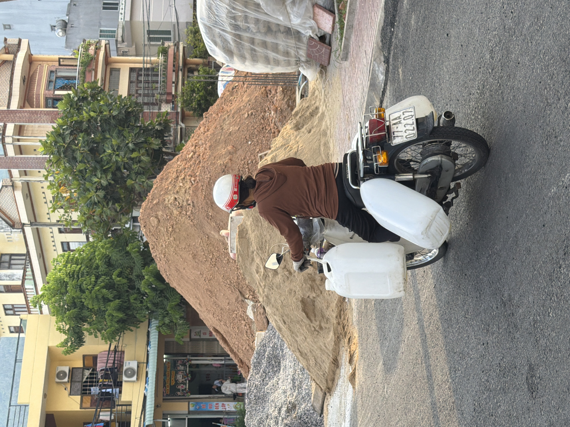 A person wearing a brown hoodie and a white and red helmet rides a Honda motorcycle on a paved road in South Quy Nhon Ward, Vietnam. The motorcycle is loaded with two large white plastic containers, one on each side. The rider is looking forward. In the background, there are piles of sand and gravel, construction materials, and multi-story buildings. The sky is visible in the upper part of the image.