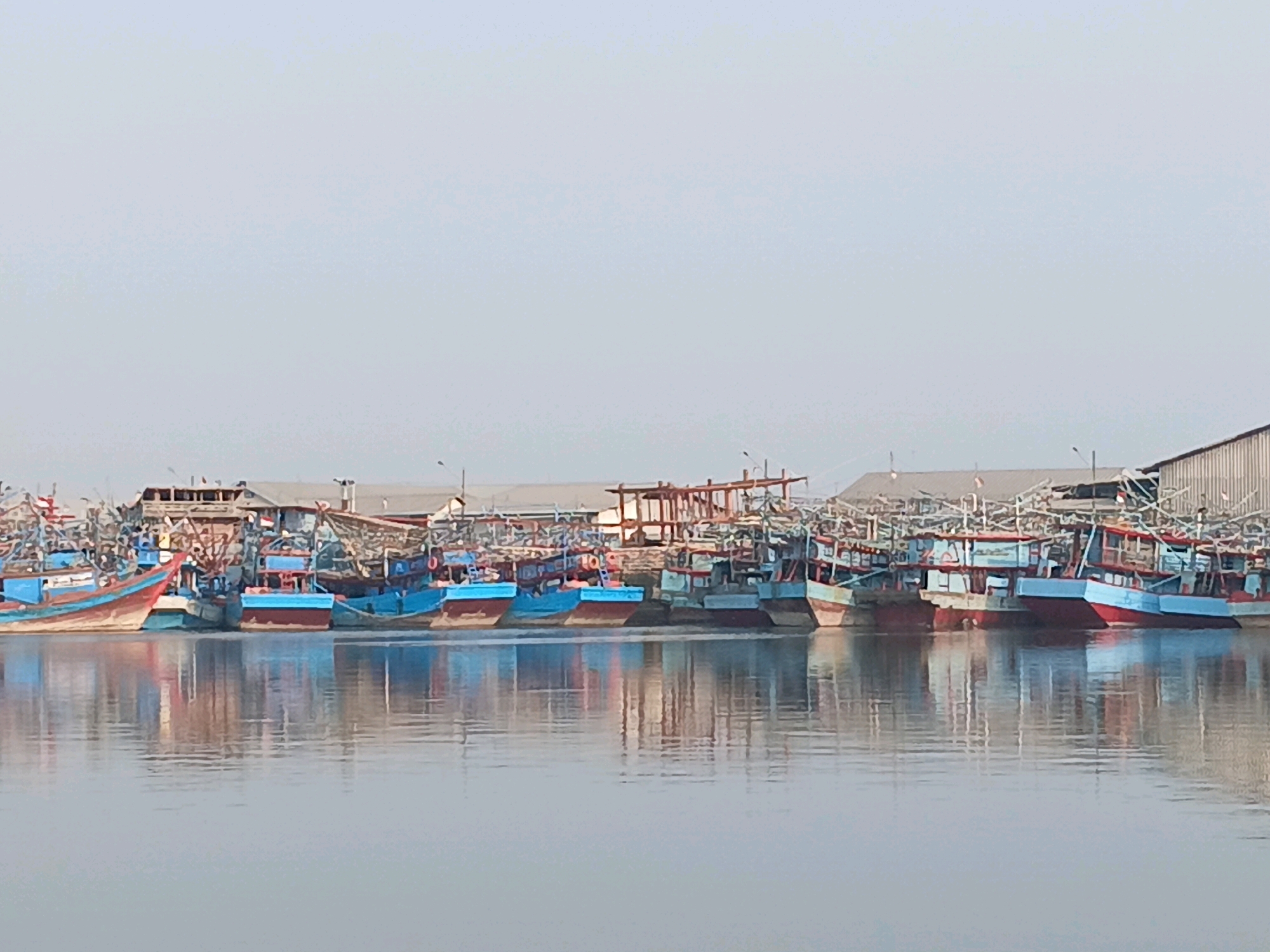 The image depicts a fleet of fishing boats docked along the shore of the city of Cirebon, Indonesia. The boats are predominantly blue and red, with some featuring white accents. They are tightly packed together, their reflections shimmering on the calm water. In the background, there are structures that appear to be warehouses or fishing industry related buildings. The sky is a pale, hazy blue, suggesting it might be a clear but slightly overcast day. The overall scene conveys a sense of a bustling fishing harbor.