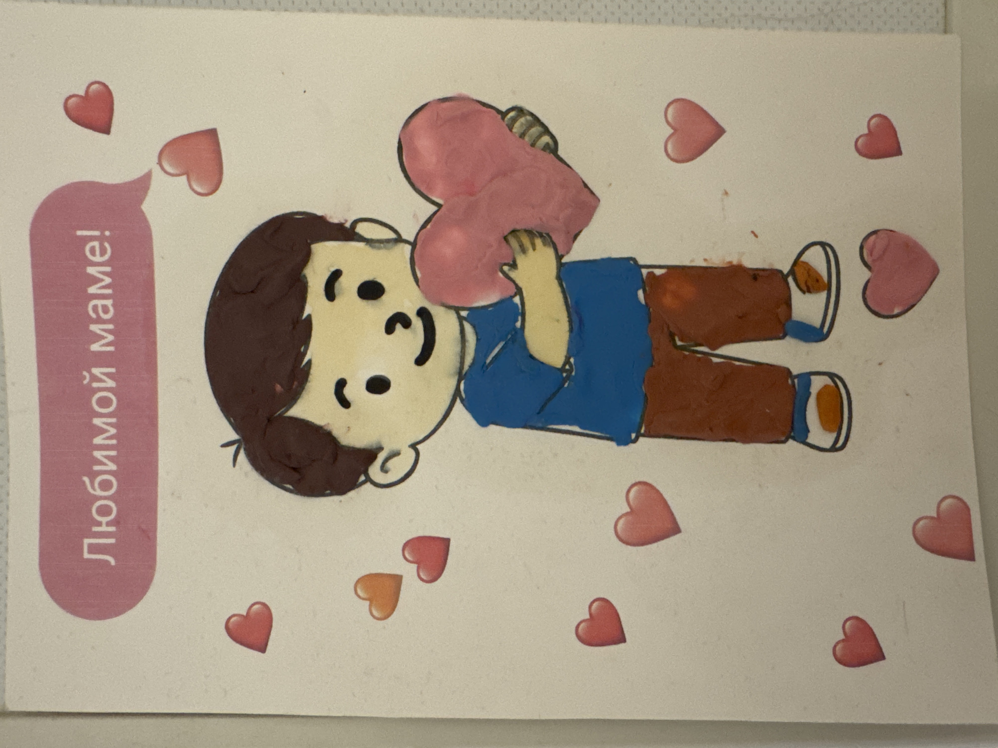 The image is a close-up, eye-level shot of a handmade card, likely for Mother's Day. The card features a drawing of a young boy made from clay or a similar textured material. He is holding a large pink heart and is surrounded by several smaller hearts in shades of pink and orange. Above the boy, within a pink speech bubble, is Russian text that reads "Любимой маме!", meaning "To my beloved mom!". The background of the card is a textured white paper. There are no contextual elements in the image to indicate a specific location in Oryol, Russia; the scene is confined to the card itself.