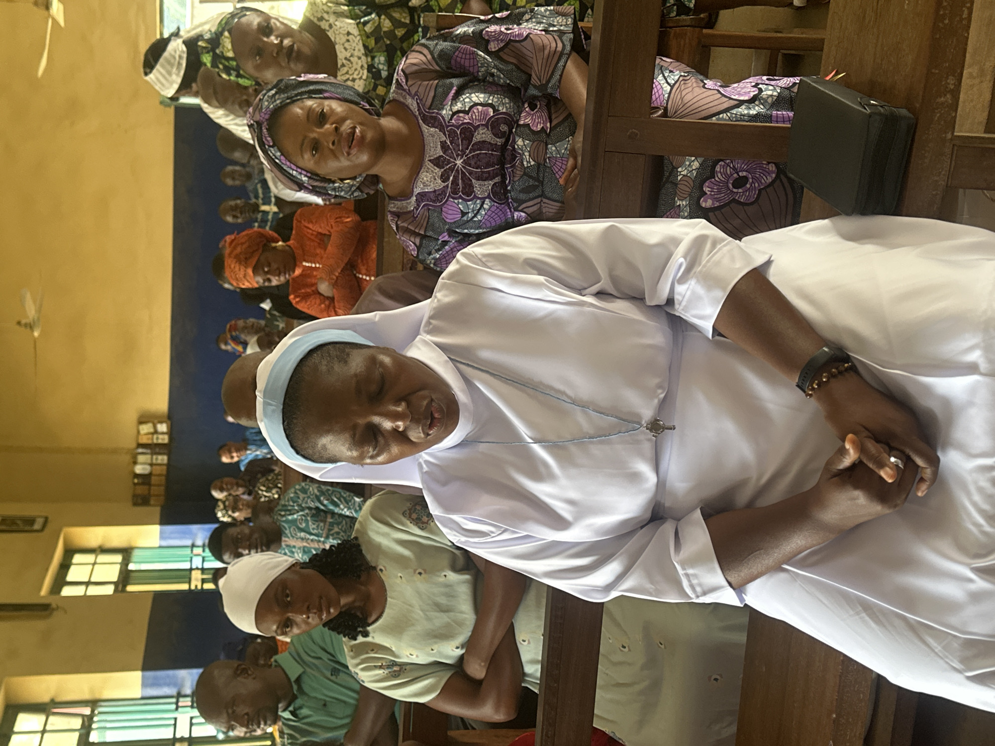In Akwanga, Nigeria, a group of people are seated on wooden pews inside what appears to be a place of worship or assembly hall. A nun in a white habit, with her eyes closed and hands clasped, is in the foreground. To her right, a woman wearing a patterned headscarf and dress looks forward, her mouth slightly open. Behind them, other individuals, including men and women, are visible, some wearing head coverings and colorful attire. The room has yellow walls and features windows with green bars. There are ceiling fans and a framed item on the wall. A small, black book lies on the pew in front of the nun.