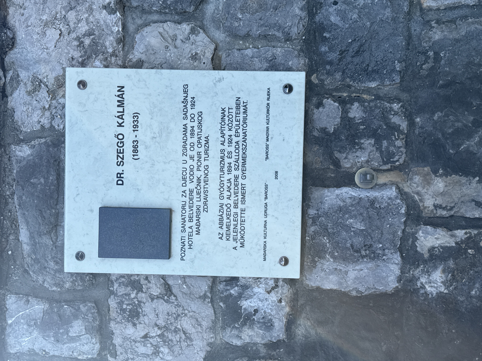The image shows a plaque mounted on a stone wall in Grad Opatija, Croatia. The plaque is light-colored with dark lettering and features a dark rectangular indentation at the top left. The text on the plaque is in Hungarian and Croatian and commemorates Dr. Szegő Kálmán (1863-1933). It mentions his role in establishing children's sanatoriums and the Belvedere hotel in Opatija. The plaque also notes that it was erected by the "MADARSKA KULTURNA UDRUGA 'BAROSS'" (Hungarian Cultural Association 'Baross') in 2008. The stone wall is made of irregular, grey stones of varying sizes.