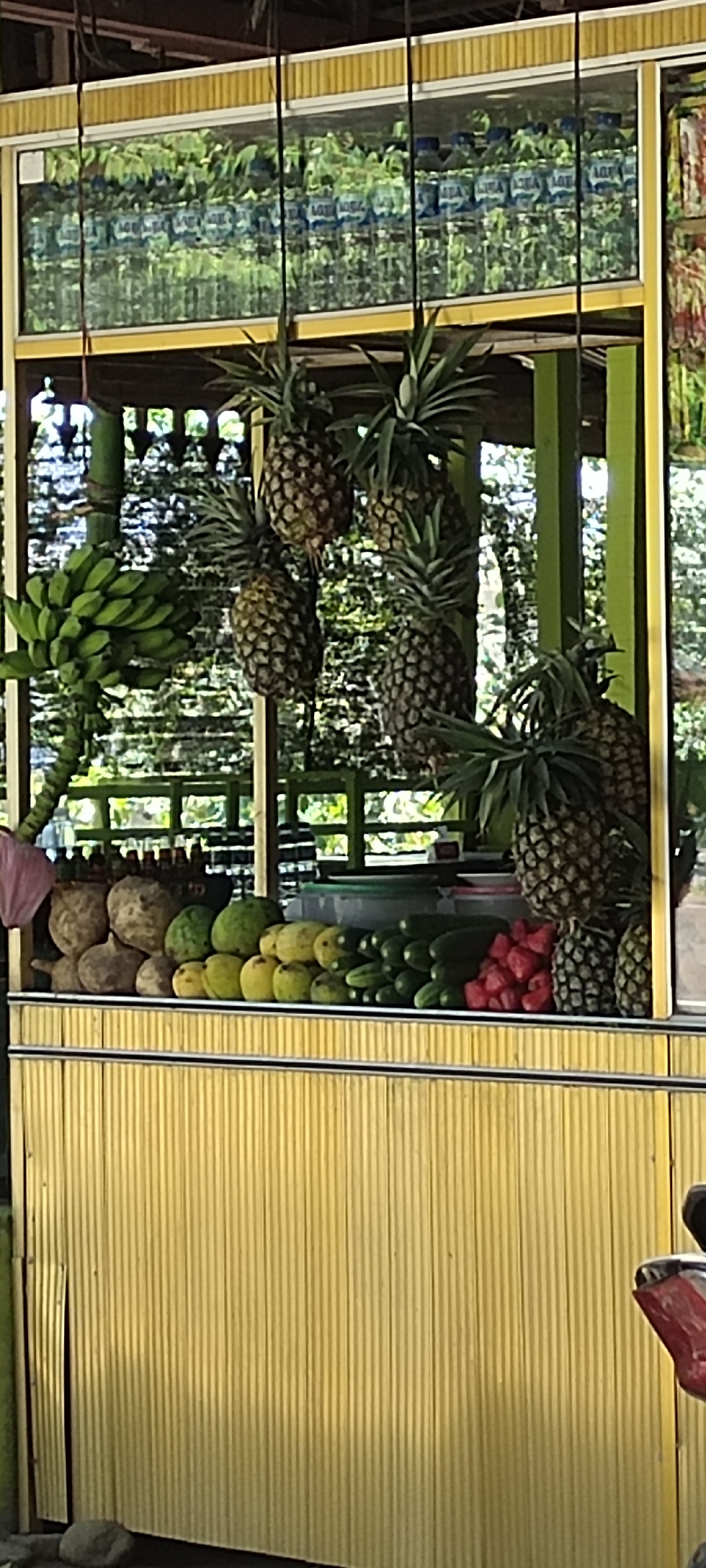 A roadside fruit stand is positioned in Lhokdagang, Indonesia. The stand displays hanging pineapples, bananas, and a variety of fruits including jackfruit, citrus fruits, cucumbers, and red salak. Bottles of water are stocked on a top shelf. The stand itself is constructed with yellow corrugated metal. A motorcycle is partially visible on the right. The background features green vegetation.