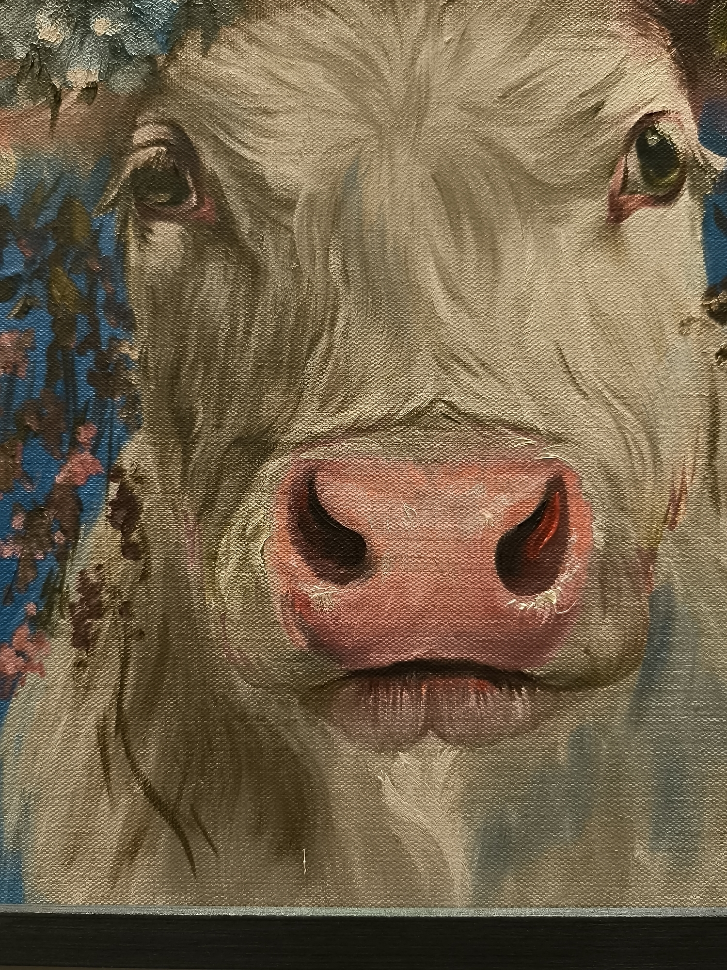 The image is a close-up painting of a cow's face, rendered in oil on canvas. The cow's fur is a light, creamy color, with visible brushstrokes creating texture. Its eyes are large and expressive, with dark pupils and reddish-pink eyelids. The most prominent feature is the cow's muzzle, which is a vibrant pink, with dark nostrils. There are hints of pink flowers and green foliage around the cow's head, suggesting it might be adorned with a floral wreath. The background is a bold blue, contrasting with the earthy tones of the cow. The painting appears to be framed by a dark border at the bottom. The provided crops highlight specific details of the cow's eyes, nose, and the textured canvas. There is no information about the city of Klaipėda, Lithuania, or any contextual details that would place the painting in a specific location.