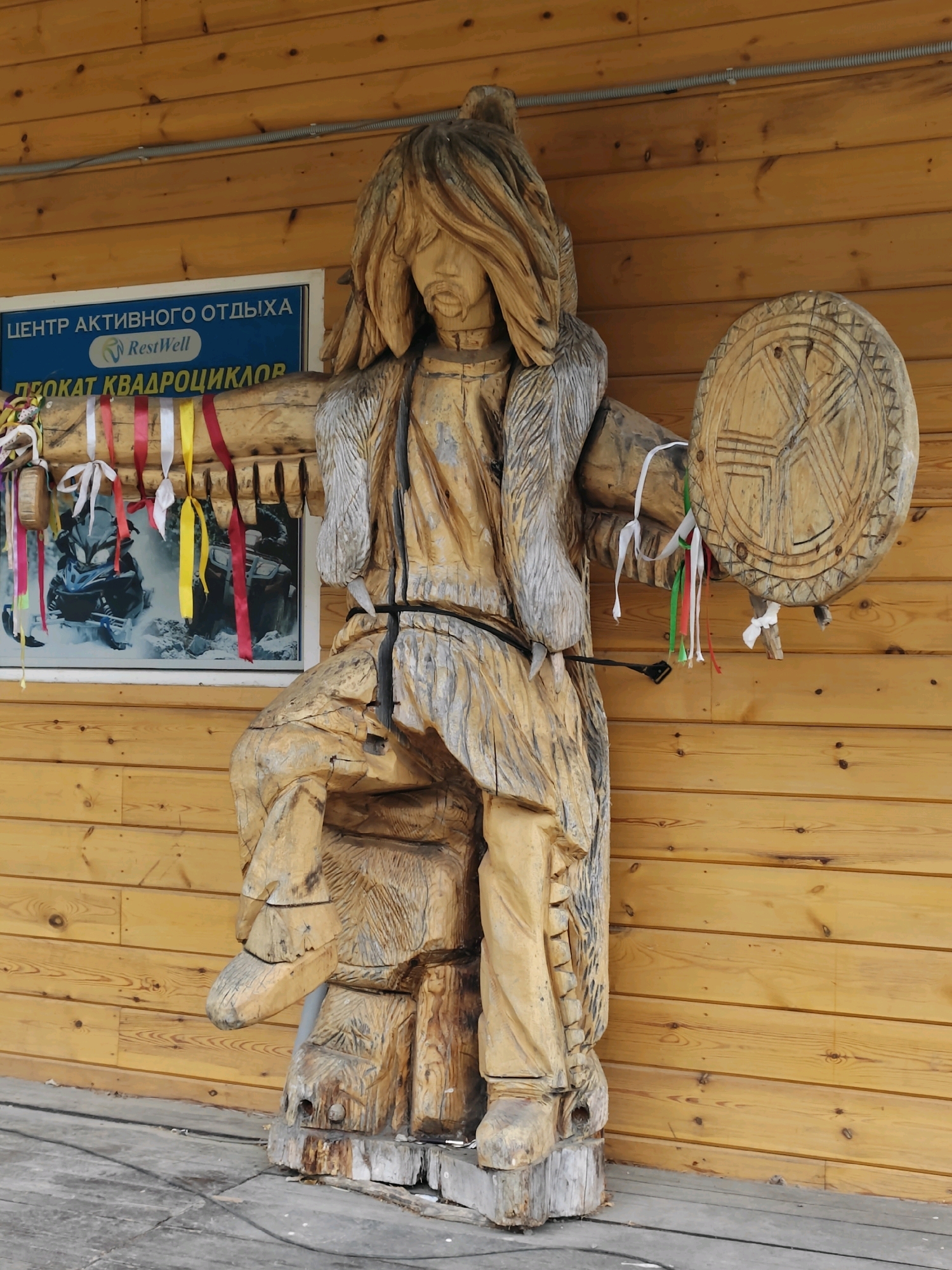 A wooden sculpture of a person is prominently displayed in front of a wooden-paneled building. The sculpture depicts a figure seated on a carved base, wearing a headdress and fur-lined garment, and holding a drum in its right hand. Colorful ribbons are attached to the drum and also hang from a wooden beam above the sculpture. Behind the sculpture, a blue sign with white and green text is visible. The sign reads "ЦЕНТР АКТИВНОГО ОТДЫХА RestWell" and below that "ПРОКАТ КВАДРОЦИКЛОВ", indicating an active recreation center and quad bike rental. A portion of a picture featuring two snowmobiles is visible behind some of the ribbons. The ground in front of the building is covered with wooden planks. The scene is located in Listvyanka (Irkutsk Oblast), Russia, a popular tourist destination on the shore of Lake Baikal.