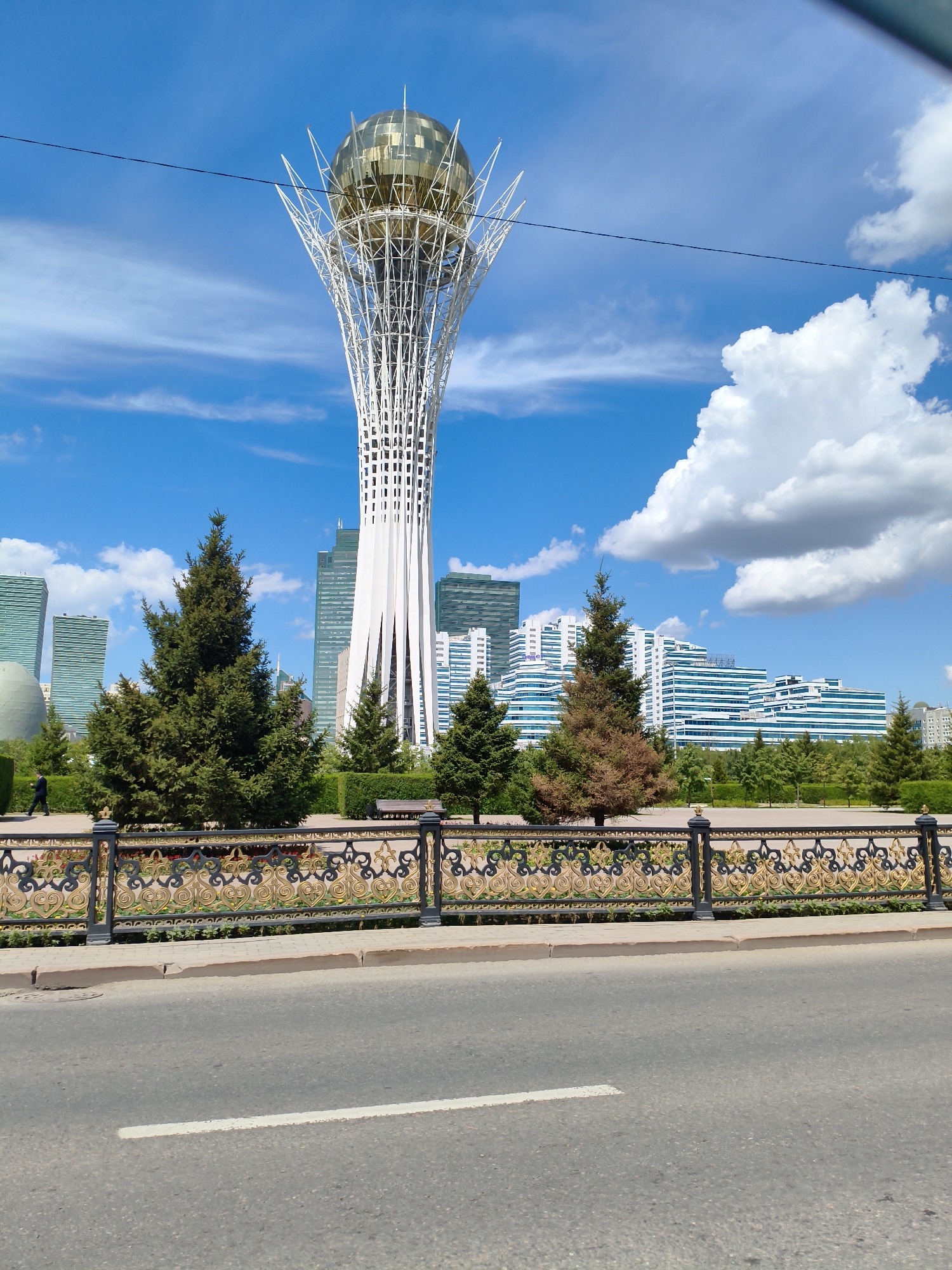 The media file captures a scenic view of Astana, Kazakhstan, featuring the iconic Bayterek Tower. The tower, a prominent landmark, stands tall with its white latticework structure topped by a golden sphere. In the foreground, a decorative black and gold metal fence lines a pedestrian walkway. Behind the fence, lush green trees and smaller conifers are planted, with a glimpse of a park bench visible.
Several modern skyscrapers with sleek, glass facades are visible in the background, creating a contrast between the futuristic architecture and the natural elements. The sky is a vibrant blue, adorned with scattered white clouds, suggesting a clear and pleasant day. The presence of a lone figure walking along the path and the overall ambiance of the scene indicate a peaceful and ordinary day in the city. The paved road in the immediate foreground with a white line painted on it suggests the image was taken from a street or roadside. The time of day appears to be midday, given the bright sunlight casting minimal shadows. The architecture and the tower itself are strong location cues for Astana, Kazakhstan. There is no visible text in the image.