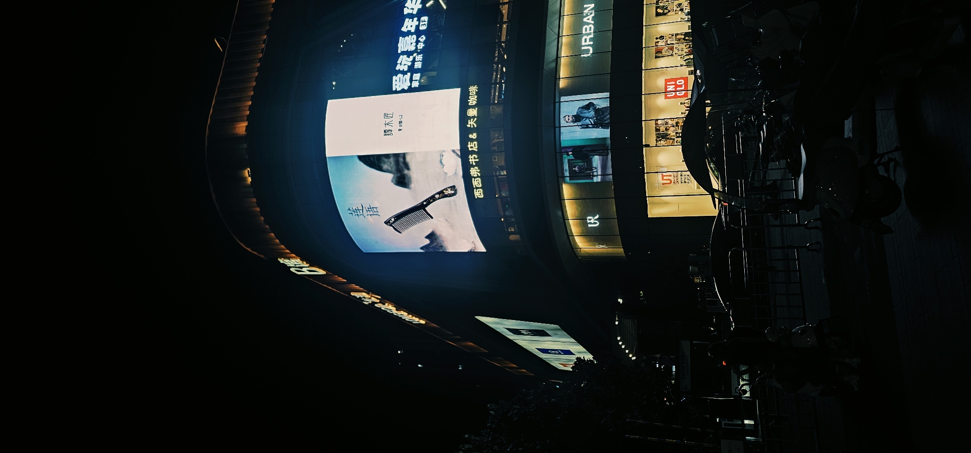 This is a night-time, low-angle, vertical shot of a modern commercial building located in the Chongqing High-tech Zone, China. The building features a large, curved, illuminated LED screen displaying advertisements. One advertisement shows a black comb with intricate floral patterns against a misty mountain backdrop and Chinese text. Another section of the screen shows text and what appears to be a logo for "Tang Mu Fen". Below the large screen, a glass-fronted retail level is visible, with neon signs for "UR" and "URBAN". Further down, the "UNIQLO" logo is prominently displayed, along with mannequins showcasing clothing. The street level is dark, with bicycles and scooters parked in front of the building, some covered by canopies. A person is seated on a scooter in the foreground. The building's upper edge is lined with warm, illuminated lights. The overall scene depicts a vibrant commercial area at night.