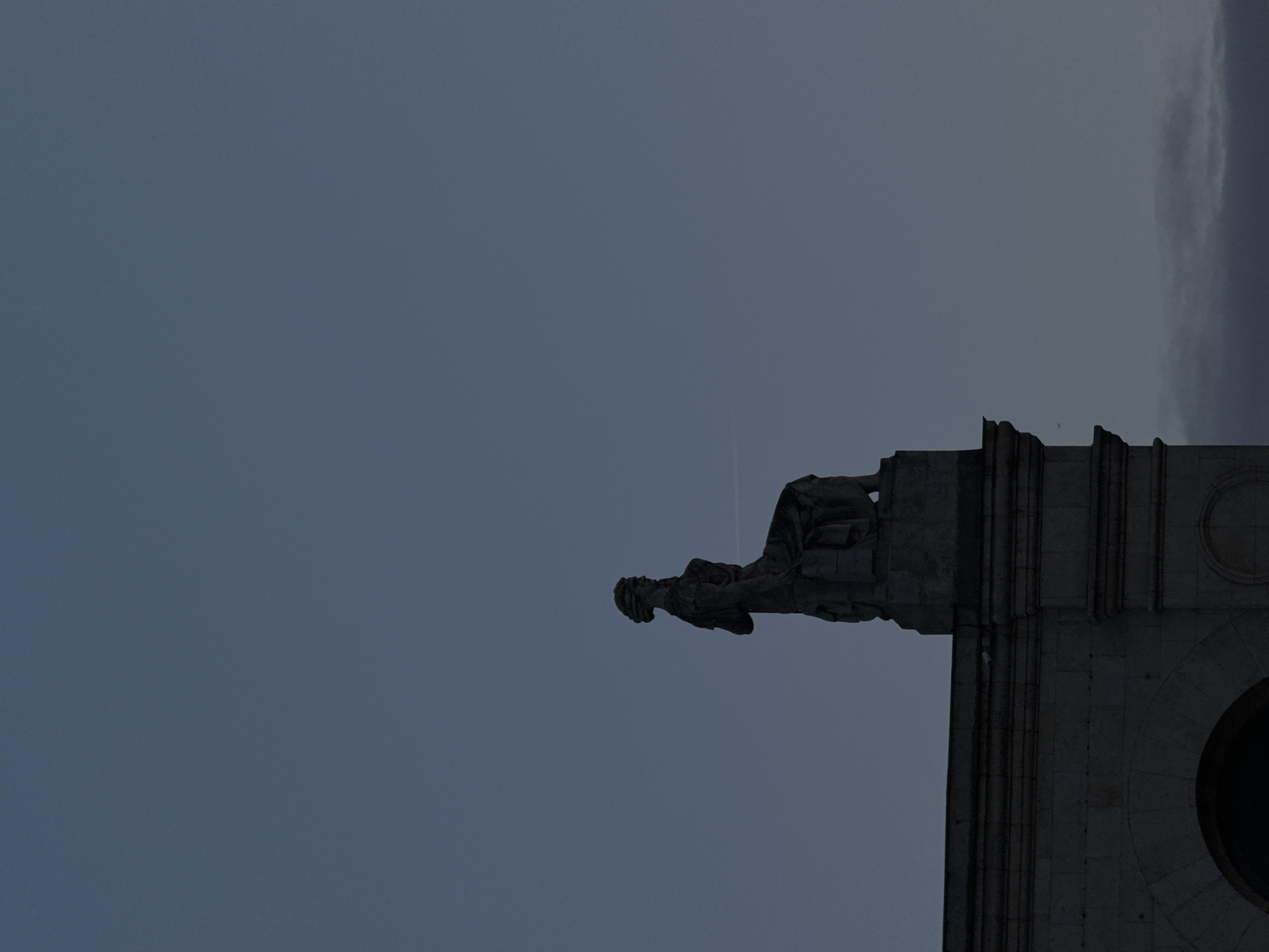 The image depicts a statue of a man atop a stone structure against a twilight sky. The statue is positioned on the right side of the frame, facing left. The man is wearing what appears to be a military uniform with a head covering. The stone structure beneath the statue is detailed with architectural features, including columns, moldings, and a circular opening. The sky is a gradient of blue, suggesting either dusk or dawn. A thin contrail stretches across the sky. The statue resembles the statue known as "Guardian Soldier" in Moscow, Russia.
