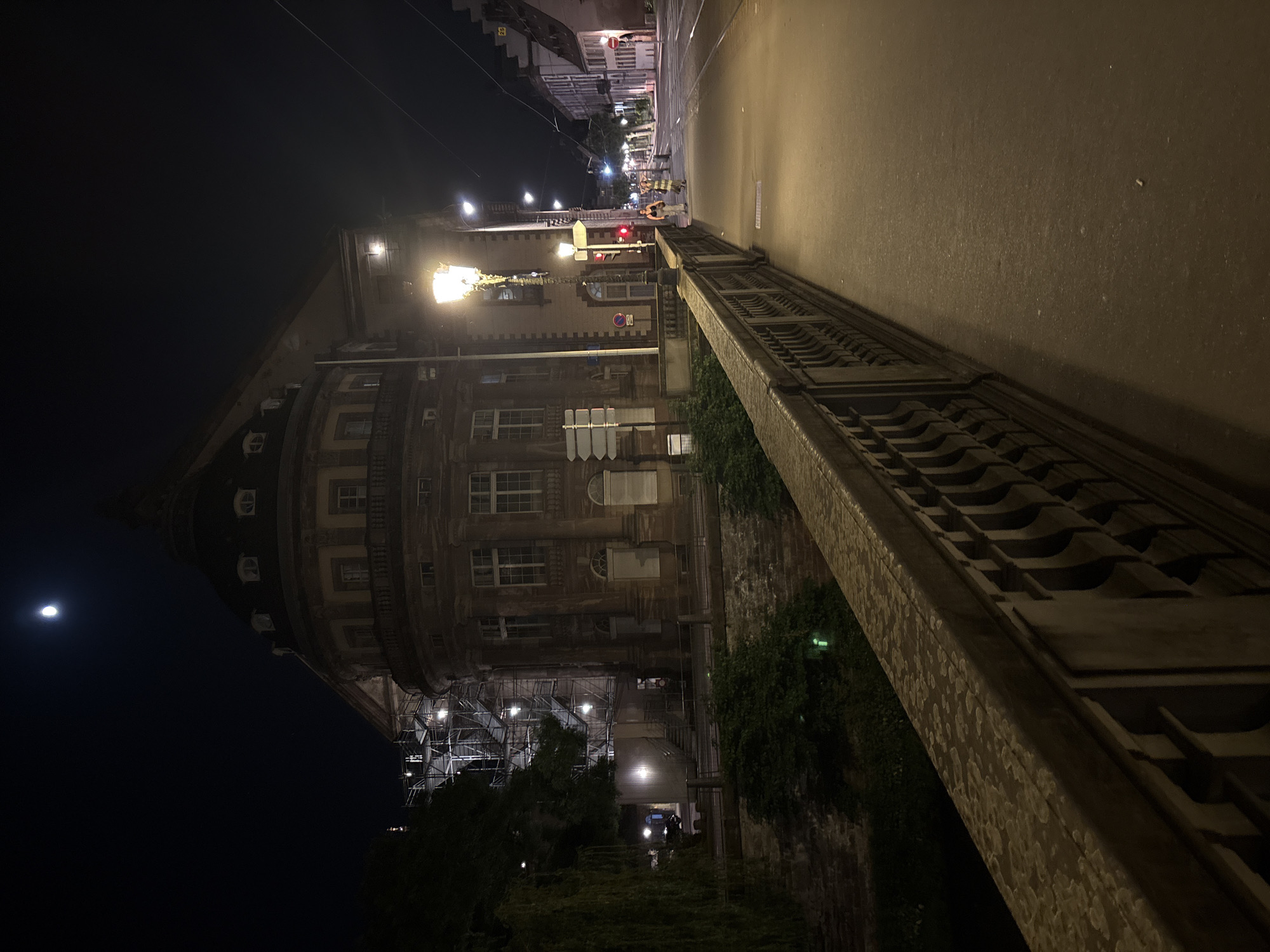 This is an angled nighttime view of the Palais de la Justice in Strasbourg, France. The building is lit by street lamps. Scaffolding obscures the lower part of the facade on the left side of the image. To the left of the building, the moon is visible in the night sky. A stone bridge runs along the right side of the image, with a road or sidewalk beyond it. Two figures appear to be working in the distance, wearing yellow and orange vests.