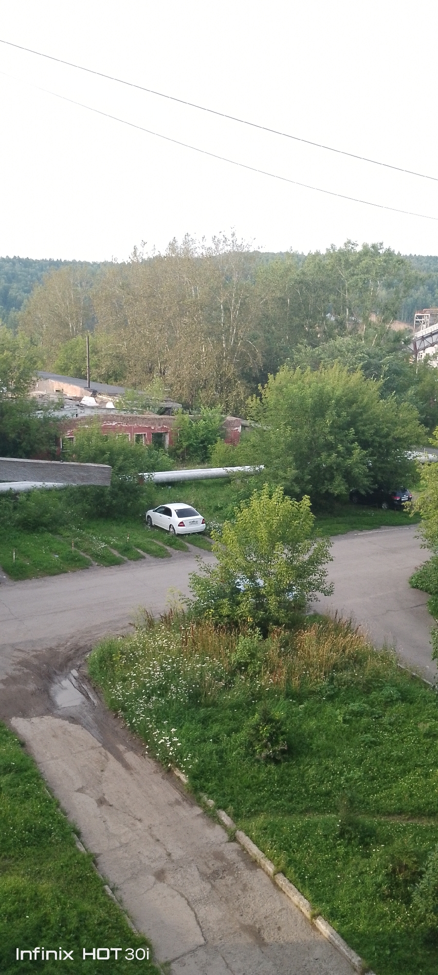 The image depicts an outdoor scene in the city of Kedrovy, Russia. In the foreground, a paved pathway, showing signs of wear and tear with cracks and some puddles, runs diagonally across the frame, bordered by lush green grass. Further into the scene, a paved road curves, with a white sedan parked to the left. A small tree with vibrant green leaves stands near the road. In the background, several buildings, some appearing dilapidated with red brickwork, are interspersed with dense green foliage and tall trees. The overall impression is that of an industrial or semi-abandoned area overgrown with nature, set against a backdrop of rolling hills covered in trees. The sky is a pale, overcast white.