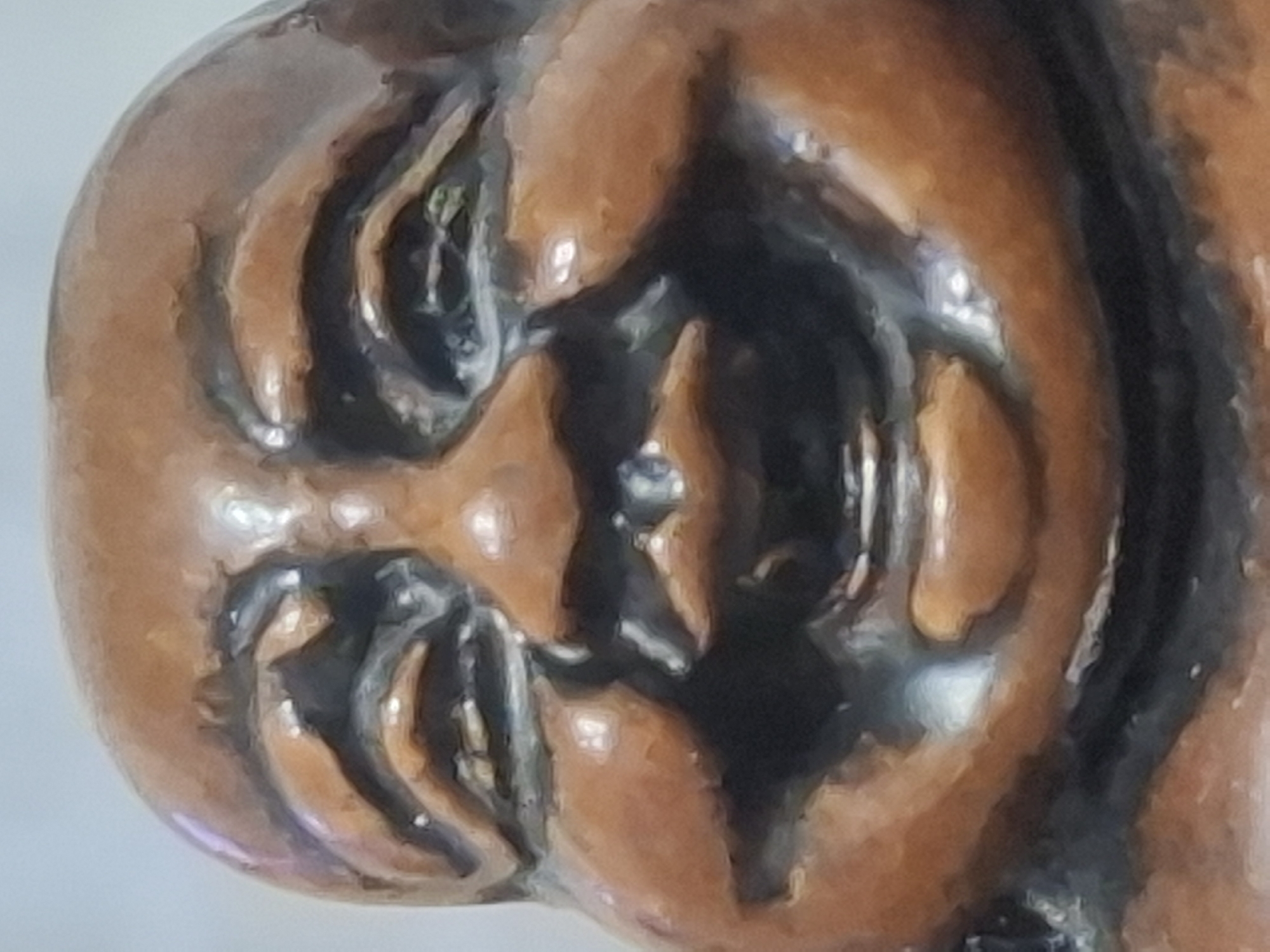 The image is a close-up of a wooden statue of a laughing Buddha. The statue is carved from a dark wood with visible grain. The figure has a round face, closed eyes with heavy lids, and a wide, open-mouthed smile revealing teeth. The cheeks are full and rounded, and the forehead is creased with wrinkles. The statue is lit from the front, casting shadows that highlight the contours of the face. The background is out of focus and appears to be a plain, light-colored surface. There is no discernible location within the city of Almaty, Kazakhstan, as the image is solely focused on the object itself and lacks any contextual environmental details.