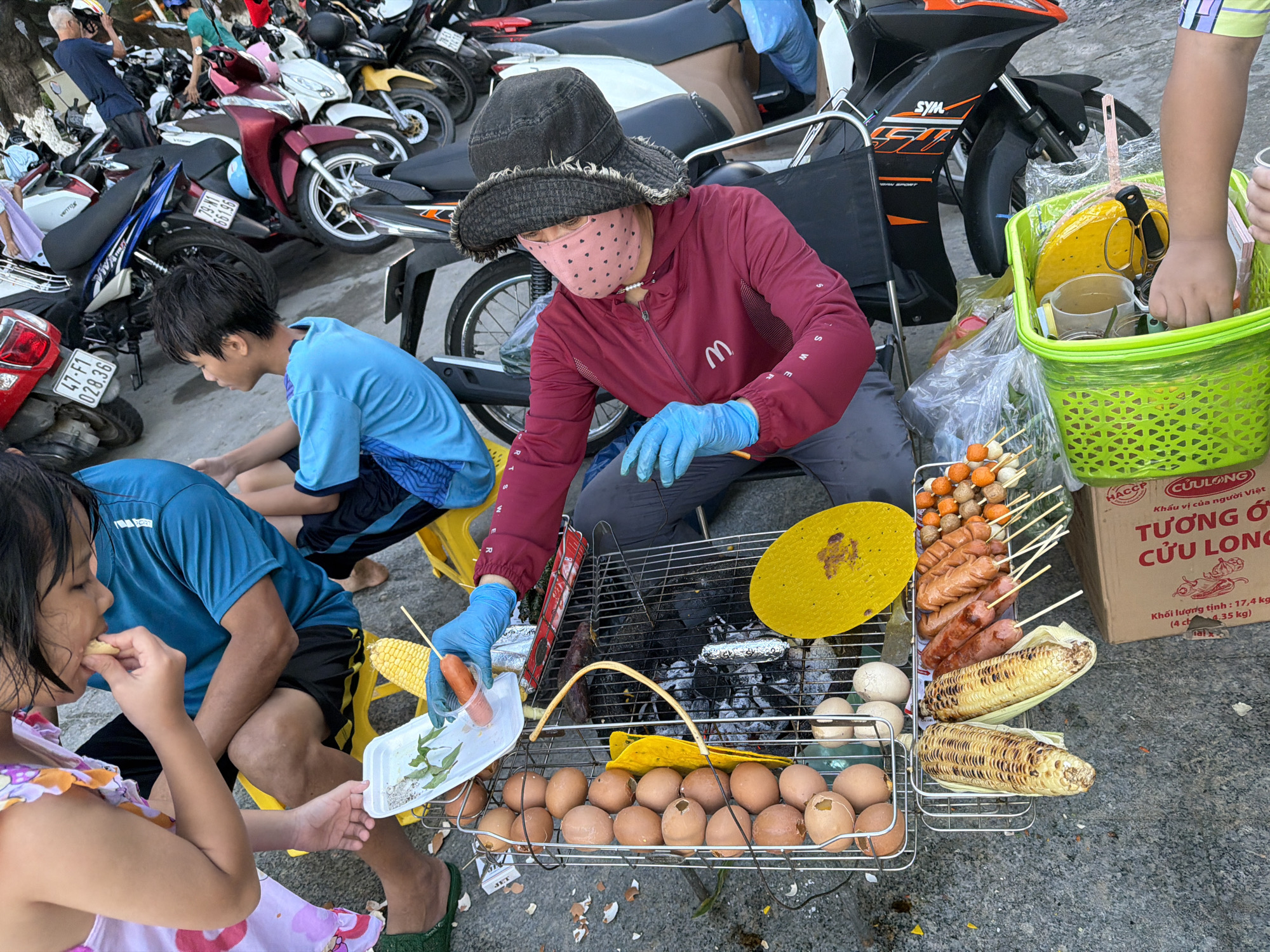 This media file captures a street food scene in Nha Trang Ward, Vietnam. The foreground features several people, including a young girl in a pink and yellow dress, who is eating. She is sitting next to a person wearing a blue t-shirt and black shorts. A person wearing a burgundy jacket, a wide-brimmed hat, and a pink face mask with polka dots is preparing food. This person is also wearing blue gloves and appears to be handling food items.
The setting is a street, as indicated by the pavement and the presence of parked motorcycles in the background. The activity centers around a portable grill where various food items are being cooked or prepared. On the grill, there are several hot dogs on skewers, grilled corn on the cob, and many eggs, some of which appear to be boiled and some cracked open. There is also a small plastic container with what looks like herbs and a hot dog on a stick being dipped into it. A bright green basket is visible, containing kitchen utensils and what seems to be a yellow frying pan.
Notable details include the various food items suggesting a meal being prepared and consumed outdoors. The presence of a face mask on one individual might indicate precautions or current health guidelines. The motorcycles in the background point to a bustling urban environment. The lighting suggests it is daytime.
Visible text includes markings on the motorcycles such as "VEITFEE" and license plate numbers. There is also text on a cardboard box in Vietnamese, which appears to be related to a food product, including "HACCP CUULONG" and "TƯƠNG Ớ CỬU LONG." Other text elements like "SYM" are also visible on the motorcycles.