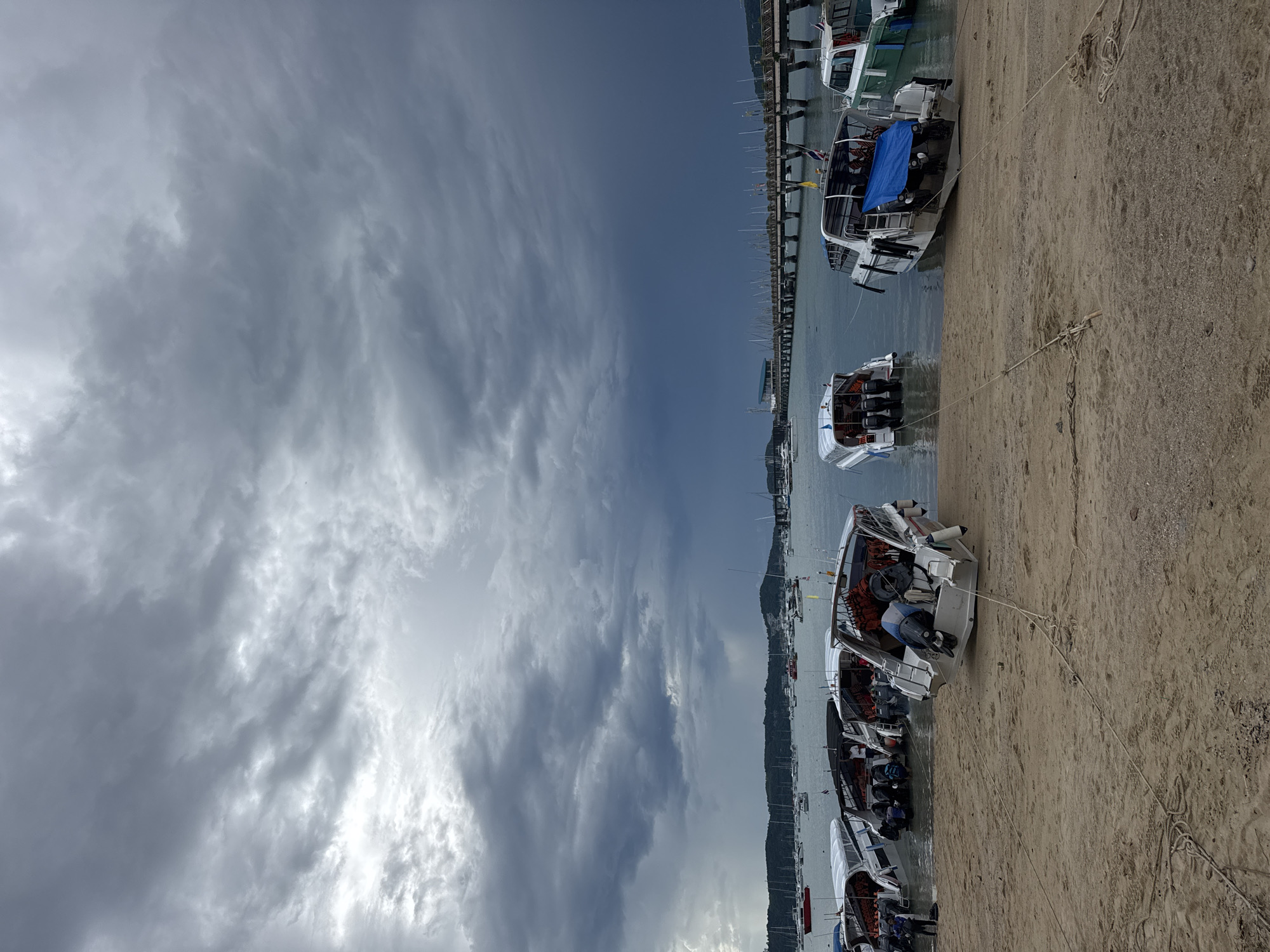 A group of speedboats are pulled up onto a sandy beach in Chalong, Thailand, under a heavily overcast sky. The boats are positioned in rows, with several of them partially submerged in the water. The sky is filled with dark, dramatic clouds, suggesting an impending storm, with a lighter, brighter area visible in the distance towards the left. In the background, a long pier extends into the ocean, lined with many boats and a building at its end. The beach itself is sandy and appears to be a public area, with ropes visible in the foreground that are likely used to secure the boats.