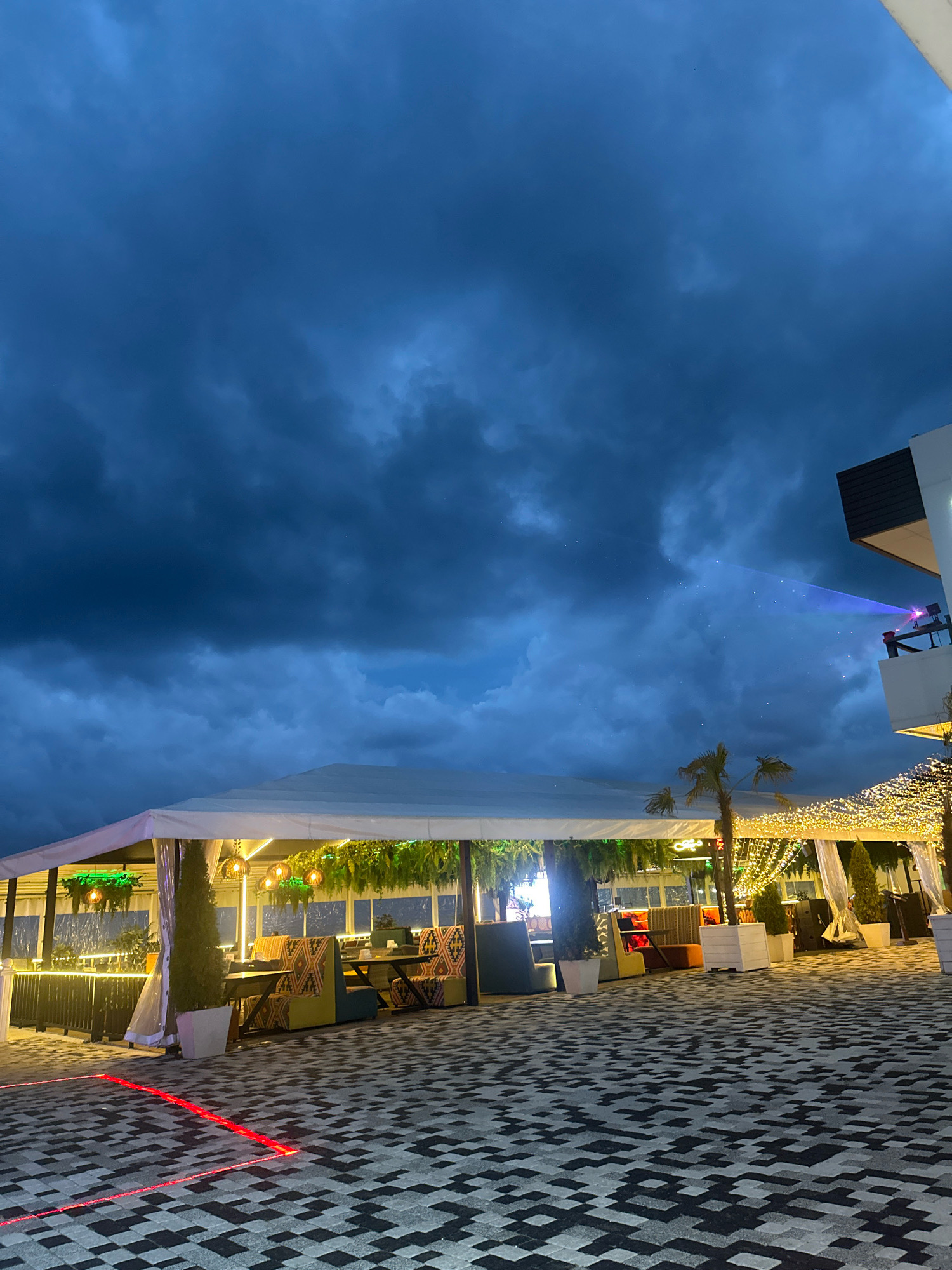 The image depicts an outdoor seating area at night, possibly a cafe or restaurant, in Lermontovo, Russia. The sky is dark and cloudy, suggesting twilight or evening. A large white tent covers a section of the seating area, which is illuminated by warm string lights and hanging lamps. Tables and chairs with patterned upholstery are visible under the tent. To the right, a modern building with a balcony is partially visible. A beam of light, possibly from a projector, cuts across the sky from the building towards the clouds. The ground is paved with a dark and light patterned tile. Red laser lines are visible on the ground in the foreground, indicating some form of demarcation or lighting. There are no people clearly visible in the scene. The overall impression is a contemporary outdoor venue set against a dramatic, cloudy sky.