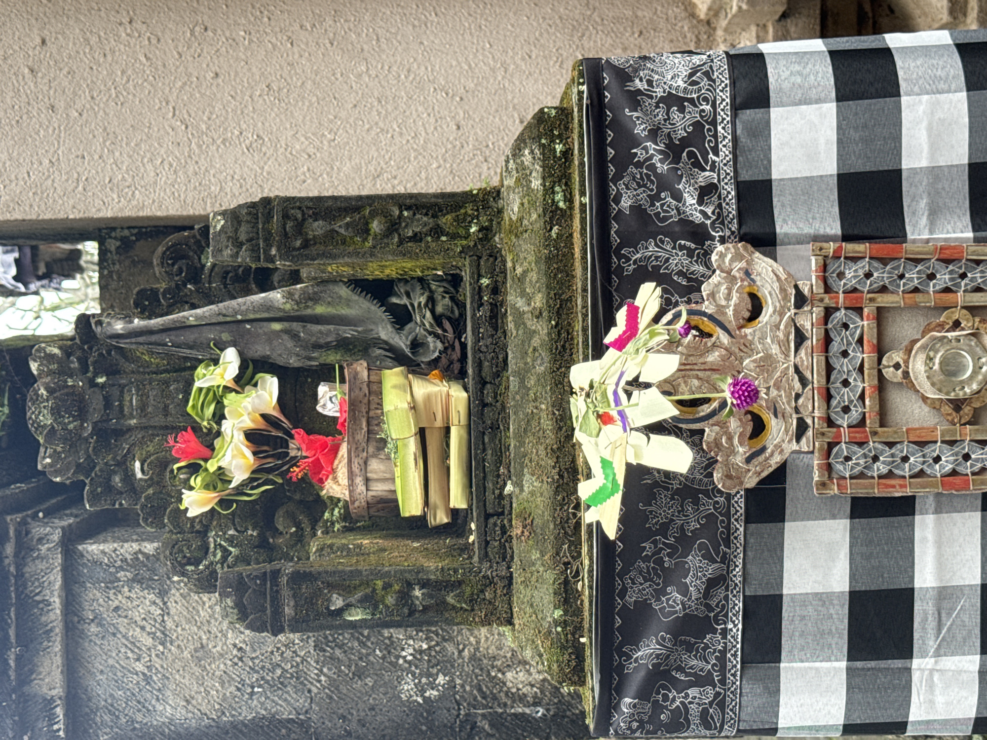 The image shows an outdoor altar in the city of Tegallalang, Indonesia. The altar is made of stone and is covered in moss. It is decorated with flowers and a black and white checkered cloth. There are also some wooden carvings on the altar. The background is a textured wall.
