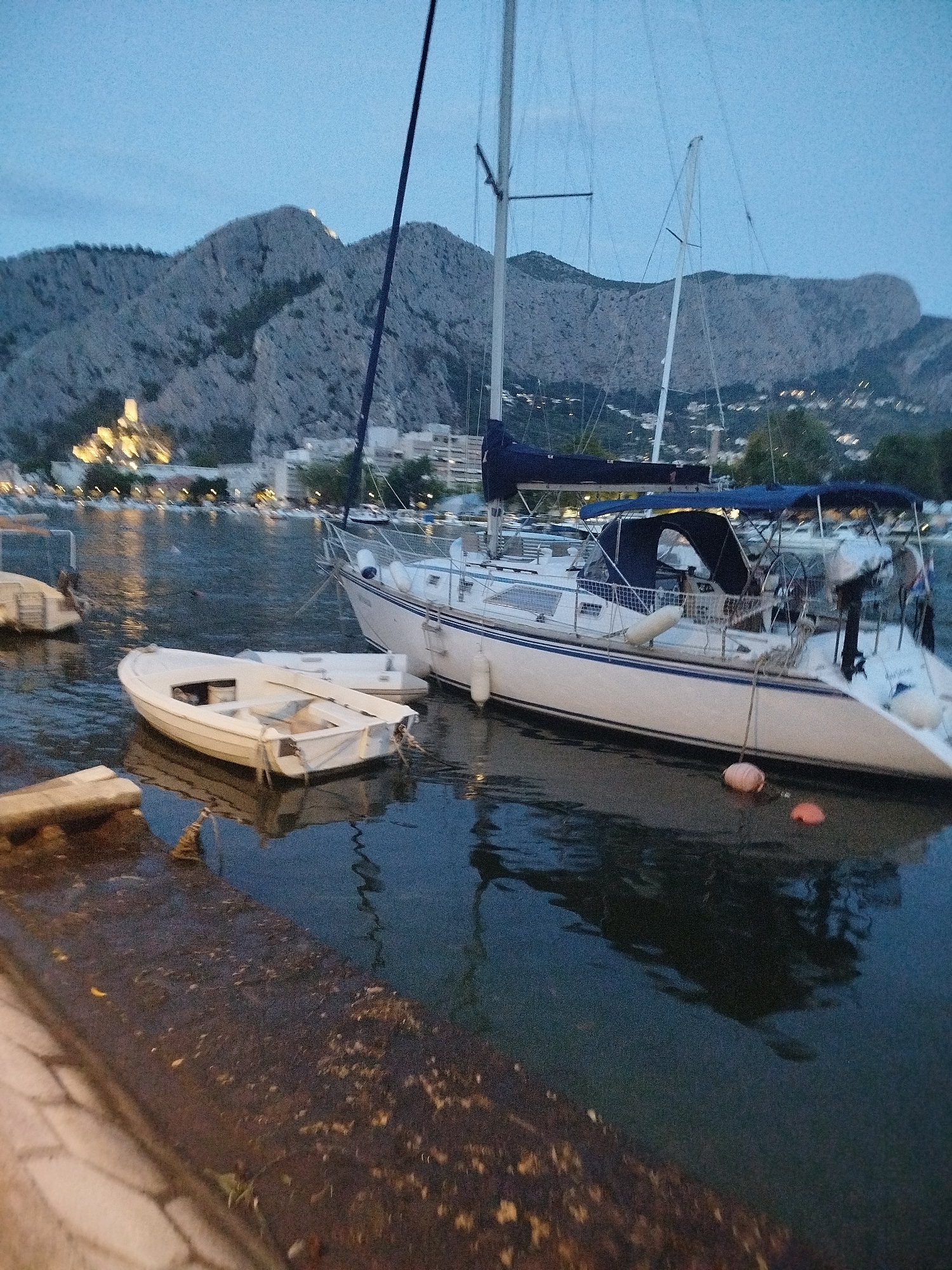 The image depicts a twilight scene in the city of Omiš, Croatia, focusing on its harbor. Two sailboats are docked in the calm, dark water. The larger sailboat, white with a blue stripe and a navy blue canopy, is in the foreground. A smaller white dinghy is moored alongside it. In the background, the distinctive, rugged mountains of Omiš rise above a town with scattered lights, suggesting a castle or fortress perched on one of the peaks. The sky is a deep blue, transitioning to a lighter shade near the horizon. The water reflects the boats and the dimly lit town. The foreground shows a concrete embankment with some debris scattered on it, and a paved walkway to the left.