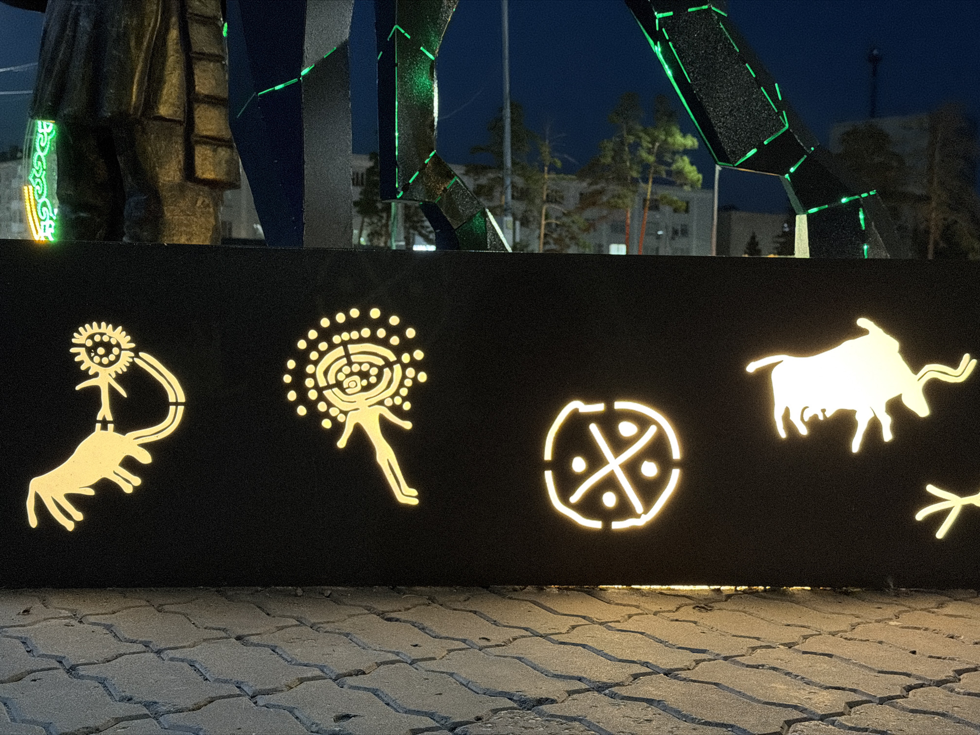 The image depicts an artistic installation at night in Pavlodar, Kazakhstan. The foreground shows a paved area with interlocking stone tiles. Behind this, a black wall is illuminated with yellow, stylized ancient petroglyphs. These include human-like figures with circular heads, a circular symbol divided into quadrants with dots, a silhouette of a bull, and a scattering of dots and lines that might represent stars or other symbols. Above the black wall, a large, dark metal sculpture is visible, with segments outlined in bright green LED lights. In the background, buildings and trees are faintly discernible against a dark blue night sky. The overall scene suggests an outdoor public art display or monument.