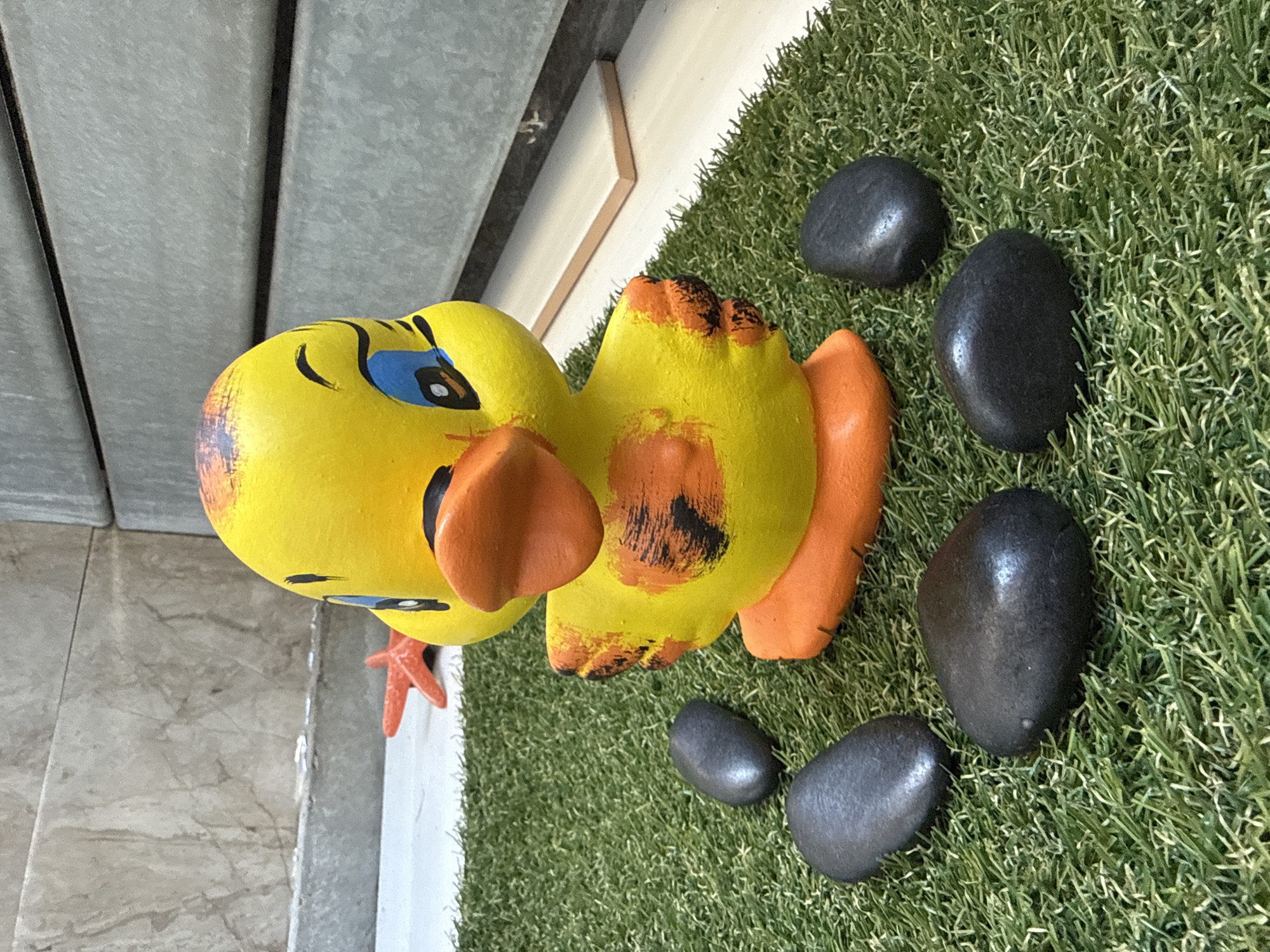 A close-up, eye-level shot focuses on a yellow ceramic duck figurine with orange feet and beak, adorned with black and orange painted markings on its wings. The duck stands on a patch of artificial green grass. Six dark, smooth stones are arranged in a circular pattern around the duck. In the background, a marble-patterned wall meets a grey, ribbed surface, with a small orange starfish figurine visible to the left of the duck. The scene is devoid of people and appears to be an outdoor decorative arrangement, possibly in a garden or patio area. The location is not specified as Sofia, Bulgaria.