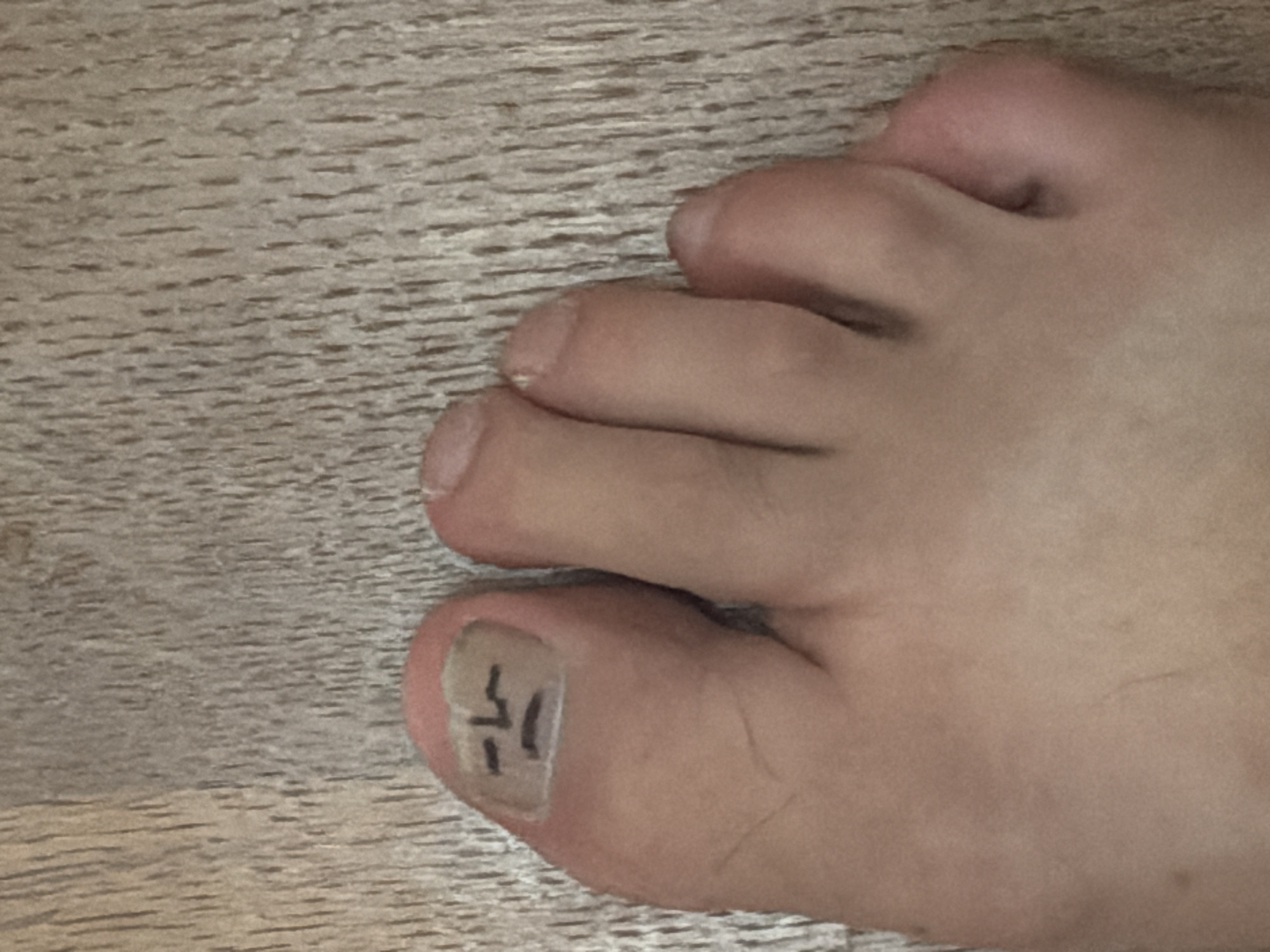 A close-up, eye-level, horizontal shot shows a person's left foot, with the toes angled slightly upward and to the right, resting on a light brown wooden floor with a visible grain pattern. The toenail of the large toe has a simple, hand-drawn face with a frowning mouth and two horizontal lines for eyes, all rendered in black marker. The surrounding skin is pale and appears to have some light blemishes. The other toes are visible to the right, with their respective toenails appearing natural. The lighting is even, and there are no shadows that would indicate the direction of the light source. The image is not associated with the city of Esch-sur-Alzette, Luxembourg.