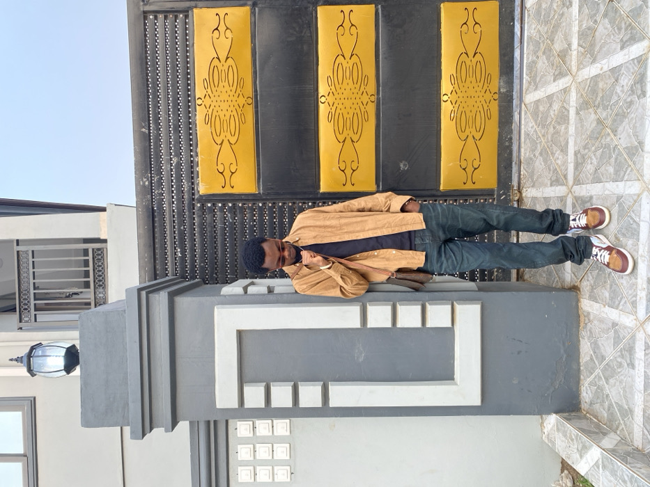 An adult male stands outdoors in the city of Joma, Ghana, positioned to the left of a large black metal gate and against a grey concrete pillar. The man, with short dark hair and wearing sunglasses, is dressed in an open tan long-sleeved shirt over a dark t-shirt, dark blue jeans, and brown, white, and maroon sneakers. A brown patterned cross-body bag hangs over his left shoulder. His left hand is raised to his chin/neck, and his right hand is in his jeans pocket, with his gaze directed downwards.
The grey pillar features white trim and is topped with an ornate dark outdoor lantern. To the right of the man is a prominent black metal gate with two central yellow rectangular panels, each decorated with black scrolled patterns. The gate's upper and lower black sections exhibit a textured or studded appearance. The ground is paved with light grey square tiles arranged in a grid pattern. In the background, a light-colored two-story building with dark window frames and a balcony with a dark railing is visible, under a clear light blue sky.