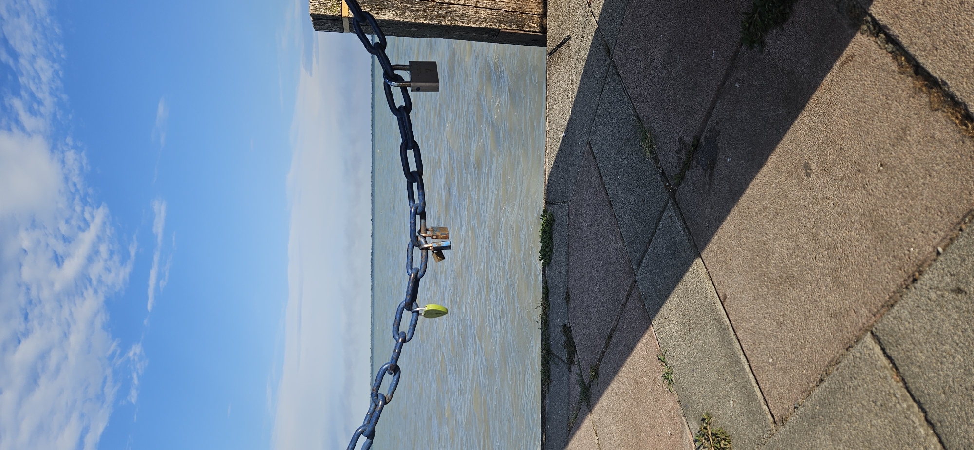 This image captures a waterside scene in Balatongyörök, Hungary. The main subjects are a thick blue chain with several padlocks attached, a body of water, and a paved walkway. The chain hangs from a wooden structure at the top left of the image, extending diagonally downwards towards the water. Three padlocks are visible: one silver padlock is near the top of the chain, another silver padlock is positioned below it, and a small green padlock is near the bottom. The water appears calm with ripples and reflects the bright sky. To the right of the water is a paved area composed of rectangular stones, with a distinct shadow cast across it, suggesting a sunny day. Some sparse green vegetation is growing between the paving stones. The sky is a clear, bright blue with scattered white clouds, indicating fair weather. The overall impression is a peaceful daytime scene, possibly a popular spot for couples to leave "love locks." The time of day appears to be late morning or early afternoon due to the long shadow. There is no visible text in the image.