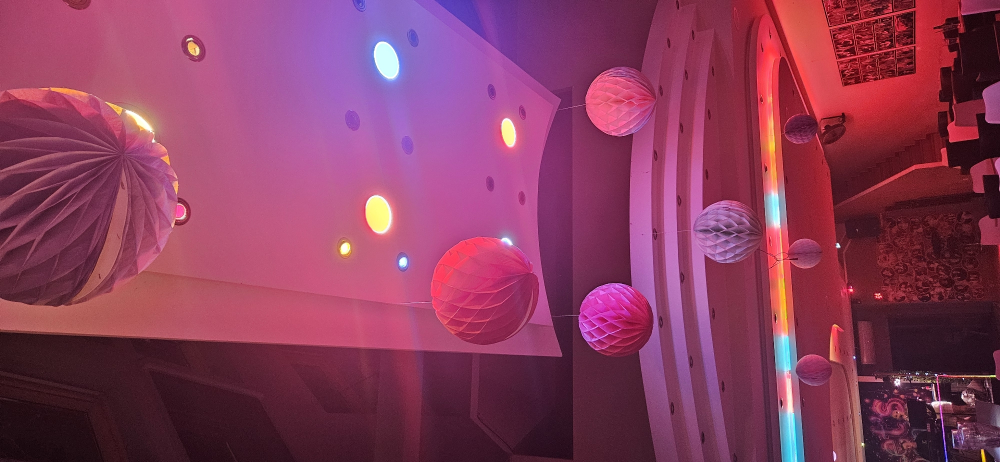 The image depicts the interior of a room, possibly a restaurant or club, in Hévíz, Hungary, illuminated by vibrant pink and red lighting. The ceiling features several circular lights, some of which are lit in yellow, blue, and white. Hanging from the ceiling are multiple decorative paper lanterns in various shades of pink and white, some appearing as spherical honeycomb designs. In the background, a DJ booth is visible with two individuals, one appearing to be a DJ. Walls are adorned with framed pictures and artwork. Stairs lead up on the right side of the room. The overall atmosphere suggests a lively and festive environment.