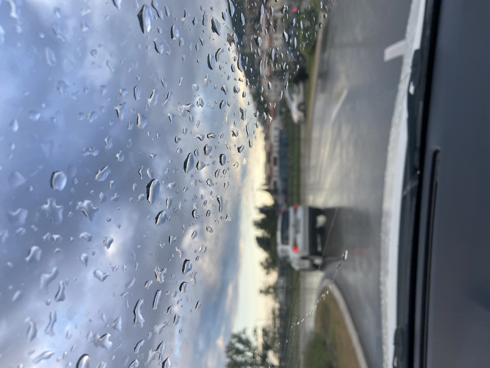 The image captures a view from inside a vehicle, looking out a window heavily covered in raindrops. The perspective is primarily focused on the water droplets, which distort and blur the outdoor scene.
The left side of the frame is dominated by numerous clear and shimmering water droplets of various sizes, clinging to the glass. Through the blurred lens of these droplets, a sky with varying shades of gray and white clouds is visible, suggesting an overcast condition or a break in the weather.
Beyond the window, an urban or suburban street scene unfolds. A light-colored car, possibly white or silver, is visible on the road in the mid-ground, appearing to be moving away or parked with its rear visible. Buildings, likely multi-story structures, and trees line the road in the background, their forms softened and indistinct due to the rain-streaked glass and depth of field. A red light, possibly from a traffic signal or building, is faintly visible in the upper right background through the raindrops. On the far right, a dark vertical section of the vehicle's interior, possibly a pillar or dashboard, and a windshield wiper blade are partially visible.
The scene depicts a moment during or shortly after rainfall in Budva, Montenegro. The lighting suggests it is either late afternoon or early evening, possibly during dusk, with soft light filtering through the clouds. There is no visible text in the image. The overall atmosphere is quiet and observant, capturing a common experience of viewing the world from a sheltered space during wet weather.