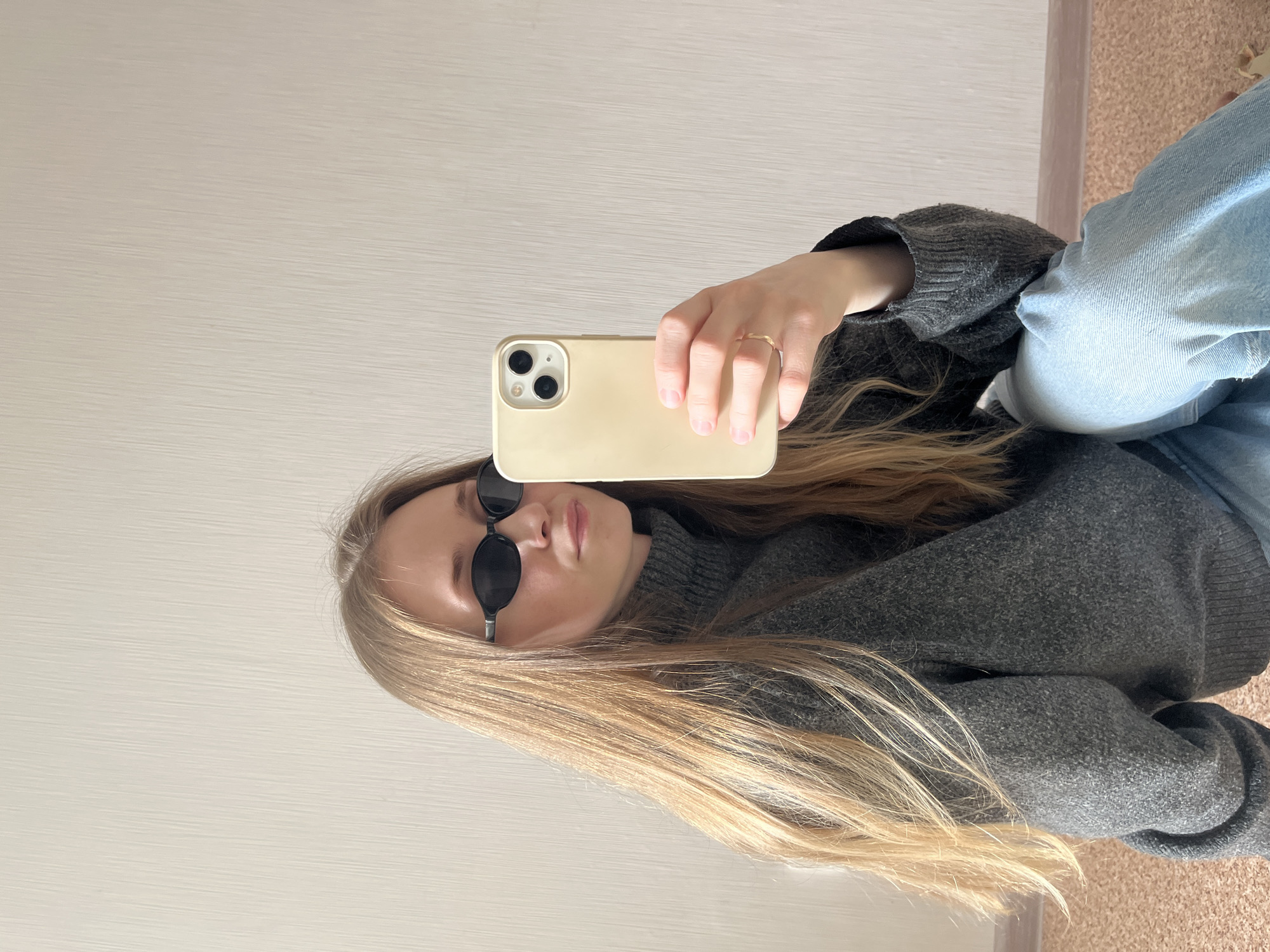 An individual, identified as a female, is depicted indoors in the city of Chaikovsky, Russia. The subject possesses long, straight, blonde hair and is attired in a dark grey turtleneck sweater, light blue denim jeans, and black oval-framed sunglasses. A thin, gold-colored ring is visible on the ring finger of her left hand. She is holding a smartphone, encased in a pale yellow cover, in her left hand, oriented towards her, indicating the act of taking a self-portrait or mirror photograph. The background consists of a plain, light-colored wall featuring a subtle vertical textured pattern. The subject is seated on a brown, cork-textured floor.