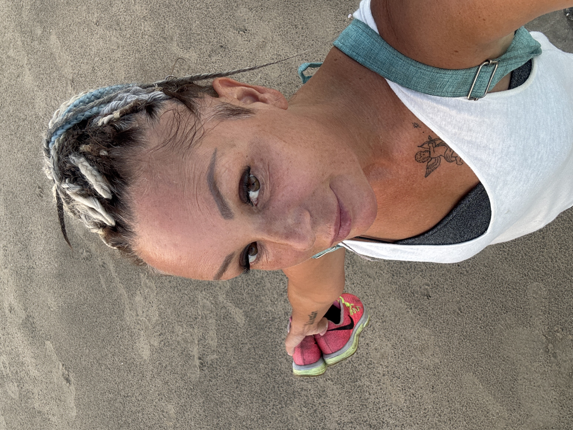 A woman stands on a sandy beach in an unknown location in Indonesia. The woman has braided hair with strands of gray and blue, and is wearing a white tank top, a gray sports bra and a teal bag strap. She holds a pair of pink and neon-green athletic shoes in her right hand. A tattoo of an angel with a bow and arrow is visible on her chest. The sand is textured.