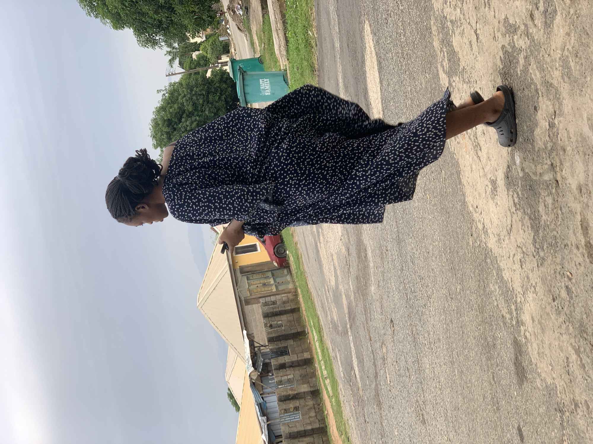 A woman with braided hair styled in a bun, wearing a dark blue dress with a white pattern, walks along a dusty, paved road. She is holding a smartphone in her right hand and looking to the left. On the left side of the road are several houses with tan roofs, some with stone walls and metal fencing. A red car is parked in front of one of the houses. Further down the road, two green trash bins are visible, one with the words "HAPPY FAMILY" printed on it. Lush green trees line the background. The sky is overcast. The scene appears to be in a residential area of Abuja, Nigeria, possibly characterized by informal settlements and limited infrastructure.