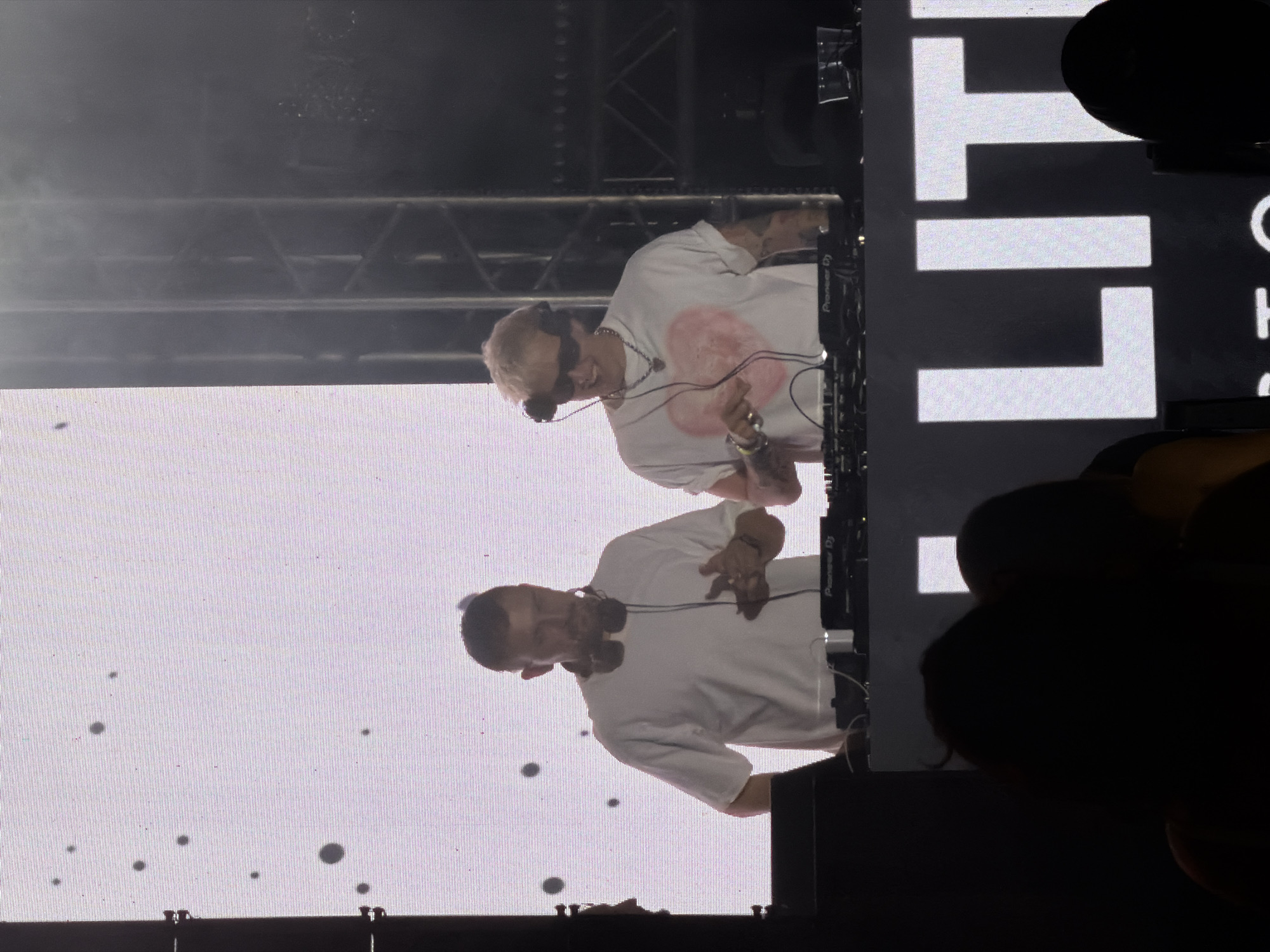 Two individuals, presumably DJs, stand behind a DJ booth on a brightly lit stage. The backdrop is a large LED screen displaying a pattern of small, dark circles on a white surface, with some horizontal lines suggesting a grid. Both individuals are wearing white t-shirts and headphones. The DJ on the left, with short dark hair, is looking down at the DJ equipment. The DJ on the right, with blonde hair and sunglasses, is smiling and looking towards the audience. The DJ booth is equipped with Pioneer DJ equipment. In the foreground, out of focus, are the silhouettes of audience members. The overall environment appears to be an outdoor music festival or concert venue, with stage lighting and truss structures visible in the background. The word "LITTLE" is prominently displayed in large white letters on a black surface in the lower portion of the image, suggesting the name of the event or artist. The image does not provide any specific information about the city of Engels, Russia.