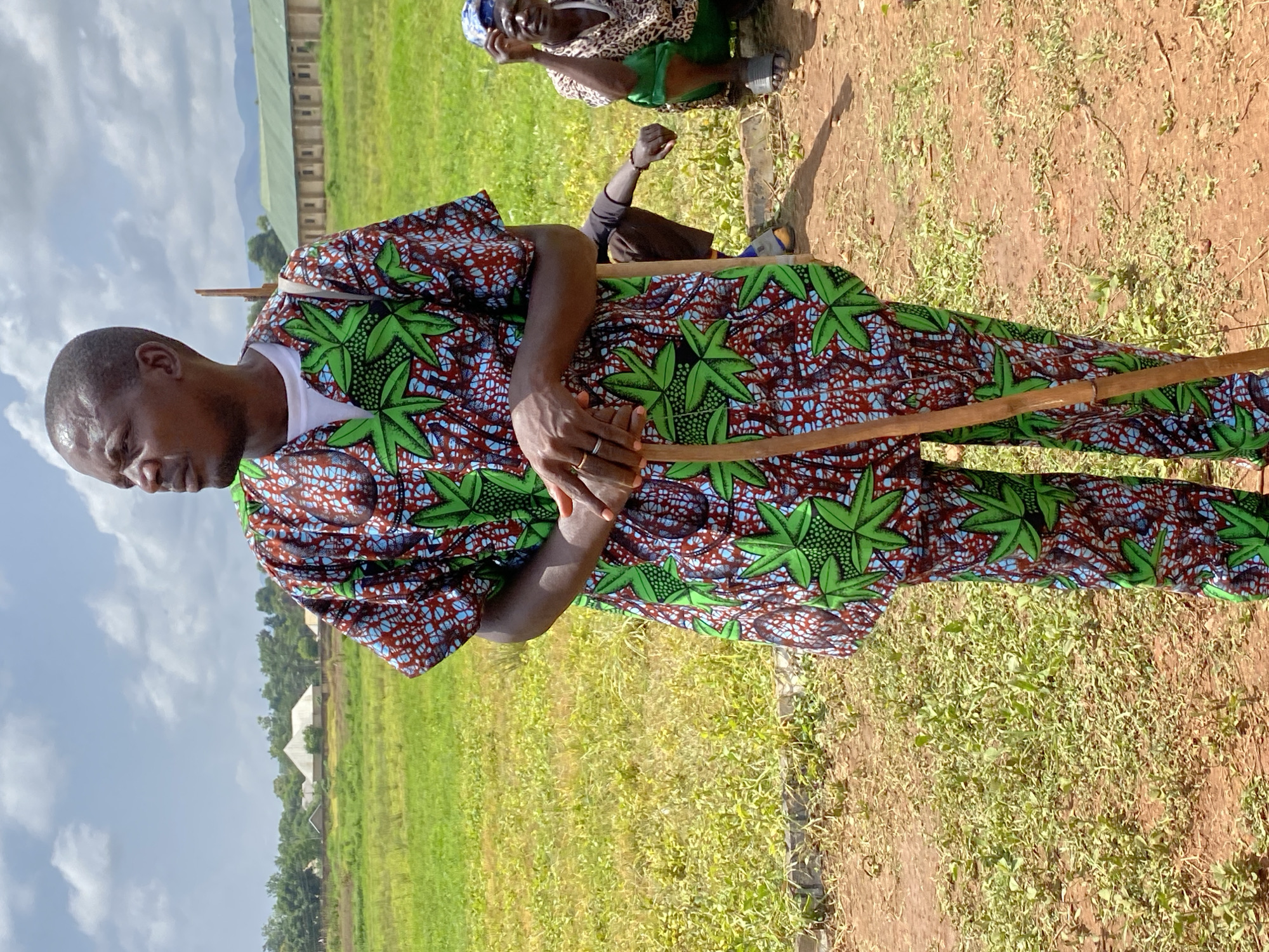 An eye-level, outdoor shot depicts a man holding a wooden stick in Maigizo, Nigeria. The man is facing to the left. He is wearing a shirt with a white under-shirt. The top shirt and matching pants are adorned with green leaf-shaped patterns and a brick-colored textured background. He is wearing a ring on his left hand. Behind him are grasses, land, buildings, and clouds. Behind the man, but slightly out of focus, are two people, one kneeling.