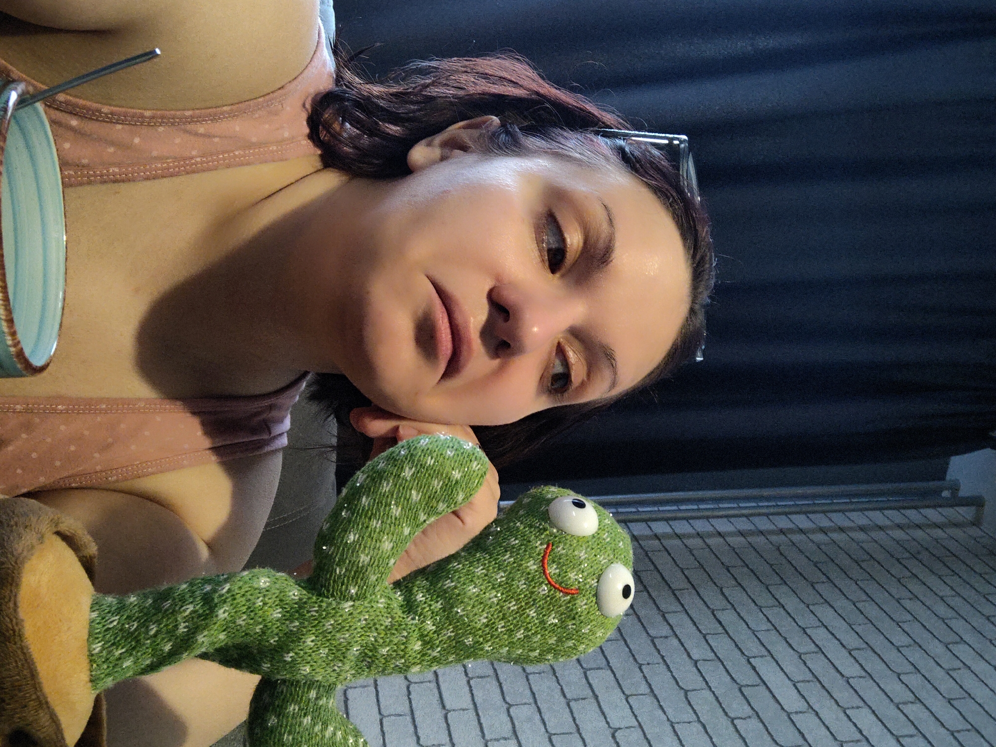 The primary subject of the image is a young adult woman with dark reddish-brown hair, seen from a slightly high, rotated perspective, appearing to be lying down. She is wearing a light pink tank top with subtle white polka dots. Her expression is neutral to slightly pensive, with her eyes cast downwards and to the side.
She holds a bright green, textured plush toy in her right hand. The toy resembles a frog or an alien, featuring large white eyes with black pupils and a red stitched smile. Small glittery specks are visible on the toy's surface.
Near her left shoulder, a light blue and brown cup containing a metallic stirring stick is partially visible. The background consists of a dark, possibly pleated or draped curtain on one side, and a light-colored, greyish-white floor with a distinct rectangular tile or brick pattern on the other. A metal rack structure is also discernible in the background.
The activity appears to be the woman relaxing indoors, holding her plush toy. The lighting is soft and ambient, characteristic of an indoor setting. There is no visible text in the image. The location context for this media file is Марковский, Russia.