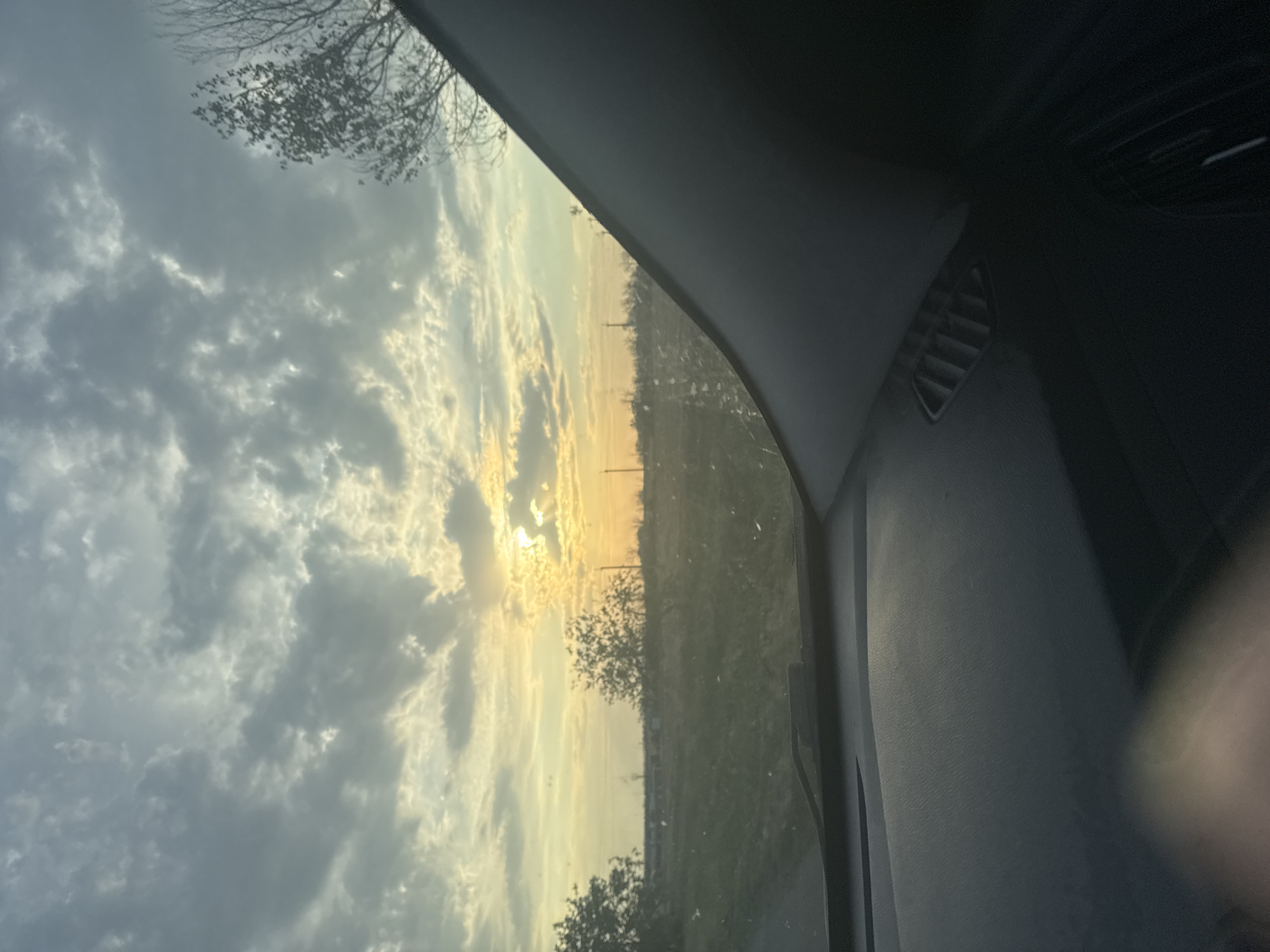 The image depicts a view from inside a vehicle, looking out through the windshield at a cloudy sky during what appears to be sunrise or sunset. The sun is visible through a gap in the clouds, casting a warm, golden light across the landscape. The foreground of the image shows the interior of the car, including the dashboard with a vent. The middle ground features a rural landscape with trees and what appear to be power lines or utility poles stretching across the horizon. The overall impression is one of a tranquil, possibly rural setting. The location is identified as Novoprimorskii, Russia.