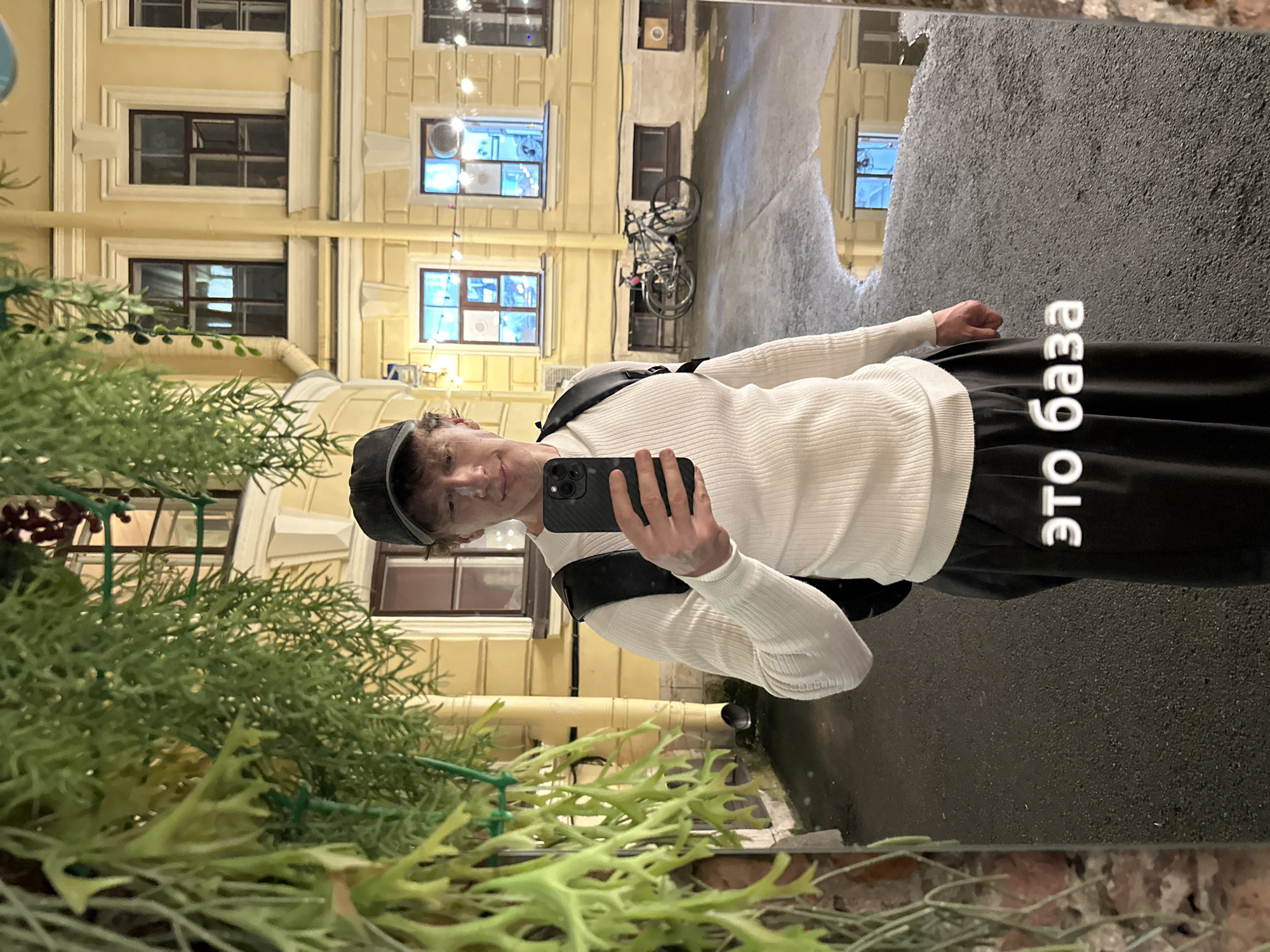 A young man with light brown hair, wearing a white ribbed sweater, black pants, and a black baseball cap, stands in an alleyway at night, taking a selfie in a full-length mirror. He is holding a smartphone in his right hand. A black backpack is visible on his shoulders. The alley is paved with asphalt and has puddles of water reflecting the surrounding buildings. To the left, lush green foliage hangs down from above. In the background, a yellow building with multiple windows is illuminated. Two bicycles are parked against the building. The text "ЭТО БАЗА" (This is the base) is superimposed in large white letters across the bottom of the image, specifically over the man's pants and the reflection on the ground. The scene is likely in Saint Petersburg, Russia, given the architectural style of the building and the Cyrillic text.