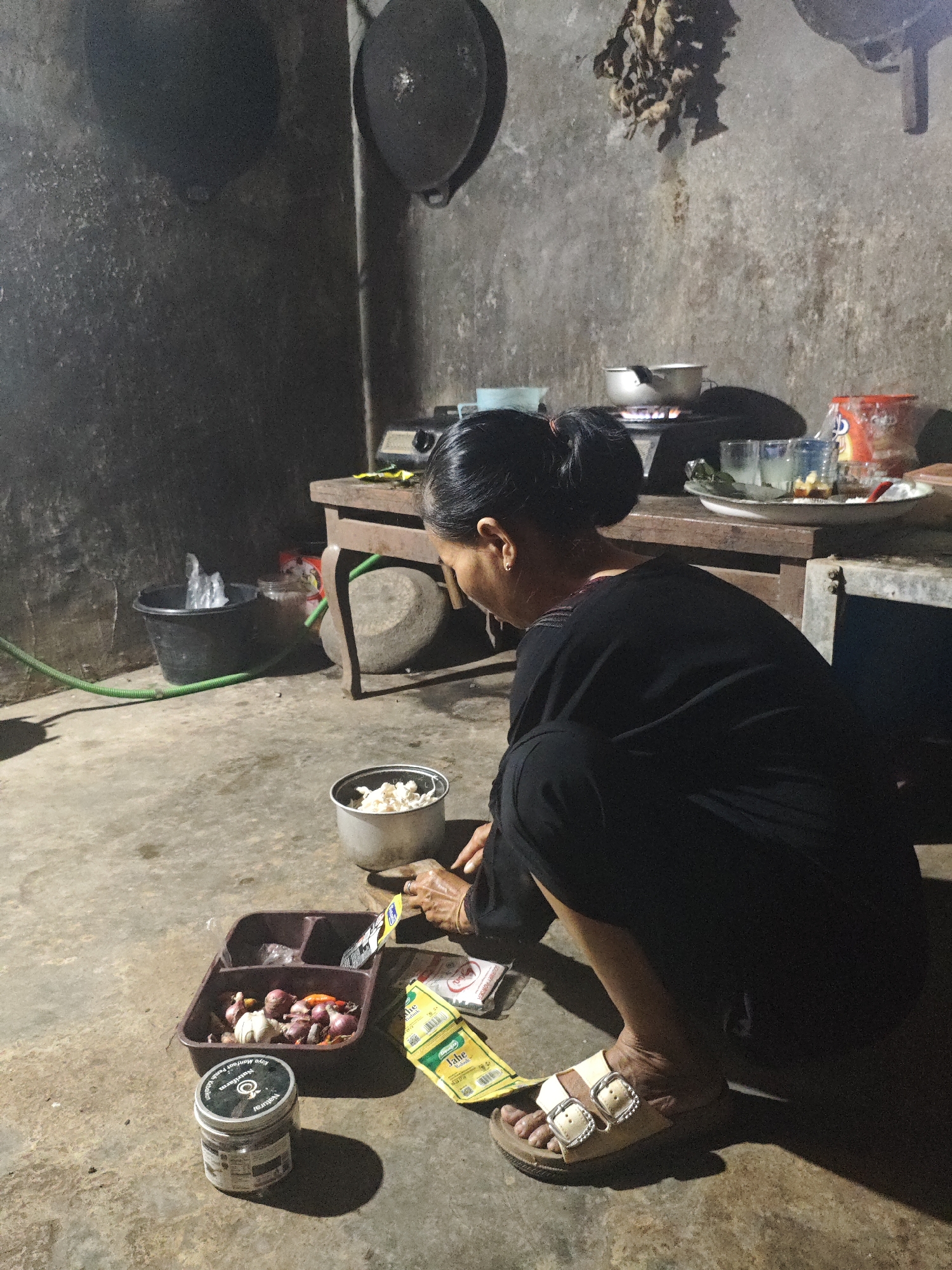 In Bantarwaru, Indonesia, a woman is seen crouching on a concrete floor in what appears to be a simple kitchen. She is wearing a black garment and light-colored sandals. In front of her, on the floor, is a brown plastic tray containing several shallots and one garlic bulb. To the left of the tray is a round, silver container. Next to the tray, to her right, are three small packets, two of which are yellow with green text. A pot containing what appears to be shredded food is situated to her right, and her hands are near it. In the background, a wooden table holds a stove with a pot on it, as well as some glasses and containers. The walls are rough and textured, and some cooking utensils are visible hanging on the wall. A black bucket with a green hose lies on the floor to the left. The lighting suggests it is nighttime or a dimly lit interior.
