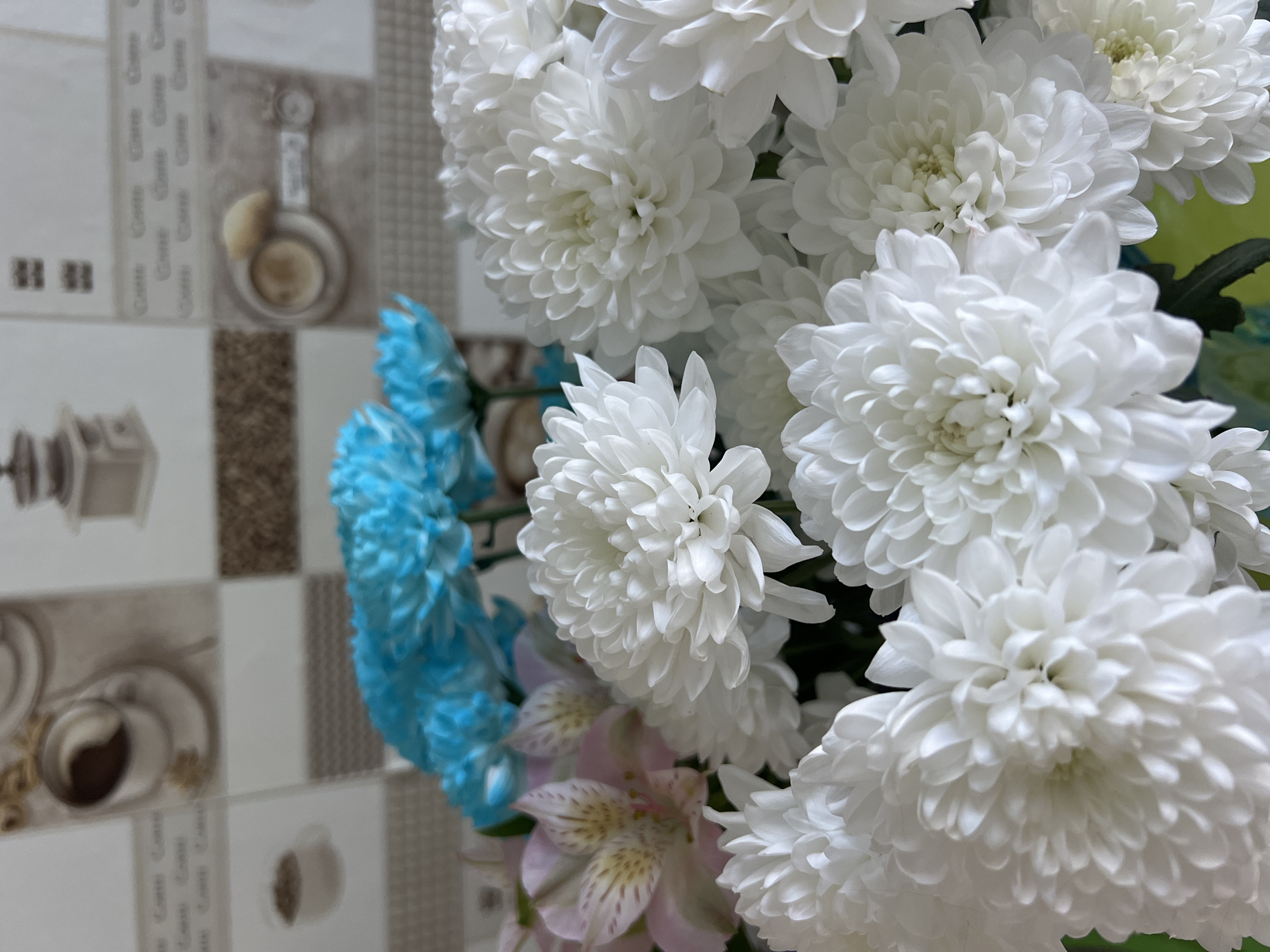 The image depicts a bouquet of flowers. The dominant flowers are white chrysanthemums, densely clustered together. There is also a smaller cluster of blue flowers and a pink alstroemeria flower visible. The background features a tiled wall with images of coffee cups and coffee-related objects. There is no indication that the image was taken in Chaikovsky, Russia.