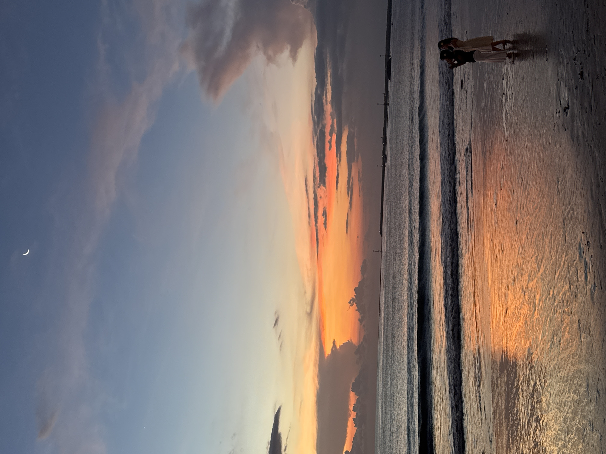 This media file captures a picturesque sunset scene on a beach, likely in Kuta, Indonesia. The sky dominates the left side of the frame (when oriented horizontally), displaying a dramatic gradient of colors. The upper portion transitions from a soft blue to a light grey, adorned with wispy, diffused clouds. Below this, vibrant hues of orange and pink streak across the sky, particularly intense towards the horizon, indicating the sun's descent. A delicate crescent moon is visible in the upper left corner of the blue sky.
The right side of the image features the calm ocean and a wide expanse of wet sand, reflecting the vivid colors of the sunset. Gentle waves roll onto the shore, creating subtle lines and patterns on the sand. In the distance, along the horizon where the sky meets the water, several faint lights or structures can be discerned.
In the foreground, standing on the wet sand near the water's edge, are two people, silhouetted against the bright reflections. They appear to be young adults or women, positioned somewhat close to each other. One person, wearing a light top and dark pants, seems to be holding a phone, possibly capturing the beautiful sunset. The other, dressed in light-colored attire, stands nearby, also facing towards the horizon. Their presence adds a sense of scale and human appreciation to the expansive natural beauty. The overall mood is serene and peaceful, capturing a tranquil moment at dusk on a tropical beach. There is no visible text in the image.