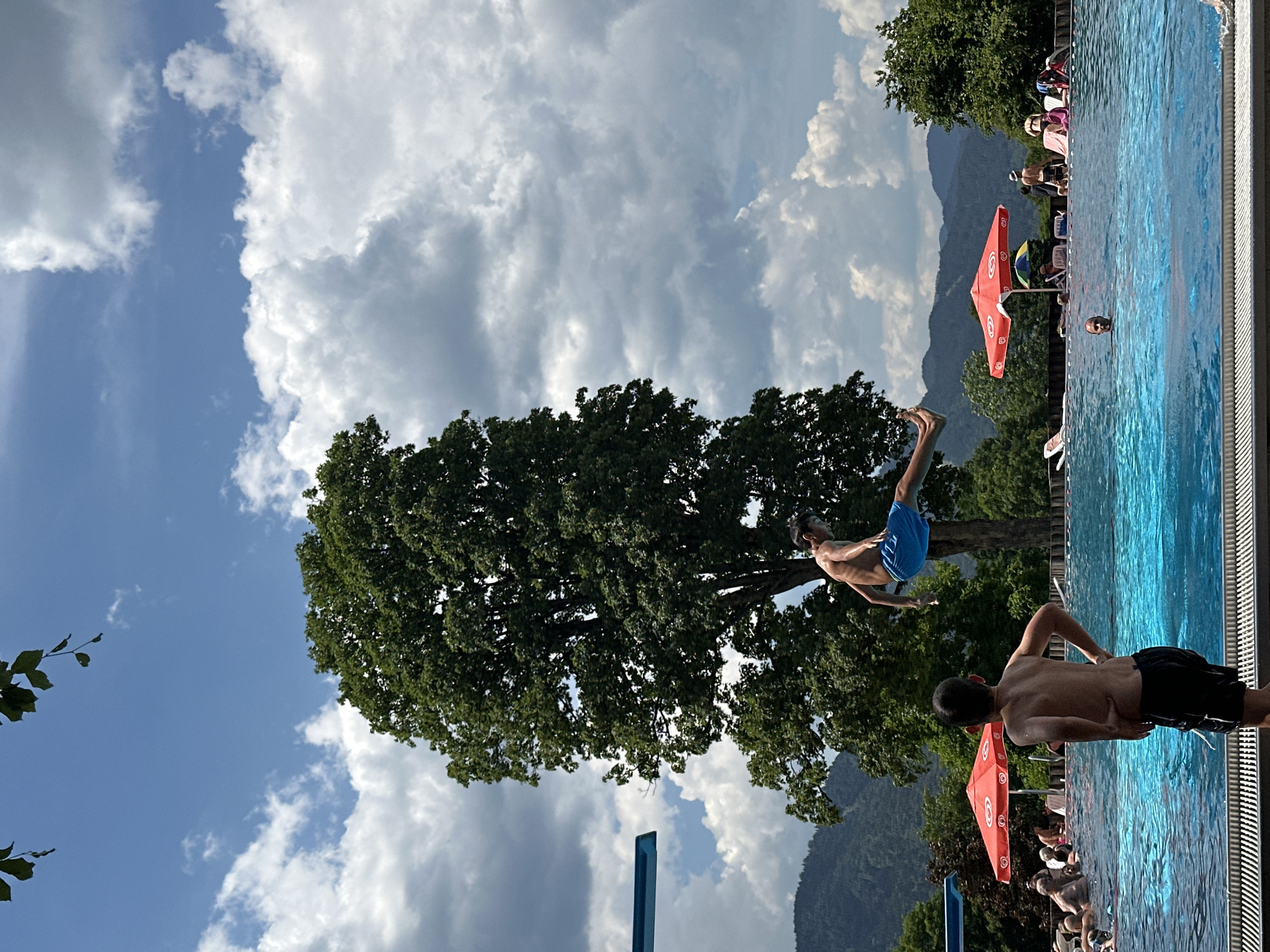 An outdoor swimming pool scene is depicted with a clear blue sky dotted with fluffy white and gray clouds. In the foreground, a young boy in black swim trunks stands facing away from the camera, his hands on his hips, looking towards the pool. To his left, another boy in blue swim trunks is captured mid-air, in the process of jumping into the pool from a diving board that is partially visible on the left edge of the frame. The pool water is a vibrant turquoise. In the background, several red umbrellas provide shade for pool-goers lounging on chairs. Lush green trees and rolling mountains form the backdrop under the dramatic sky. The overall impression is a lively summer day at a public swimming facility. The scene is located at the Oberammergau swimming pool in Oberammergau, Germany.